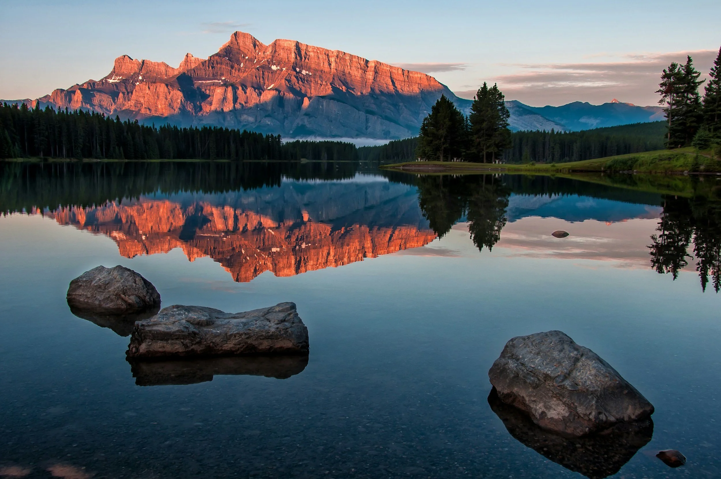 Sunrise over a calm lake with large rocks in the foreground, surrounded by pine trees, with a mountain range in the background reflected in the water.