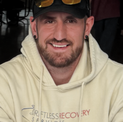Close-up of a smiling man with a beard, wearing a black cap and a cream hoodie, indoors.