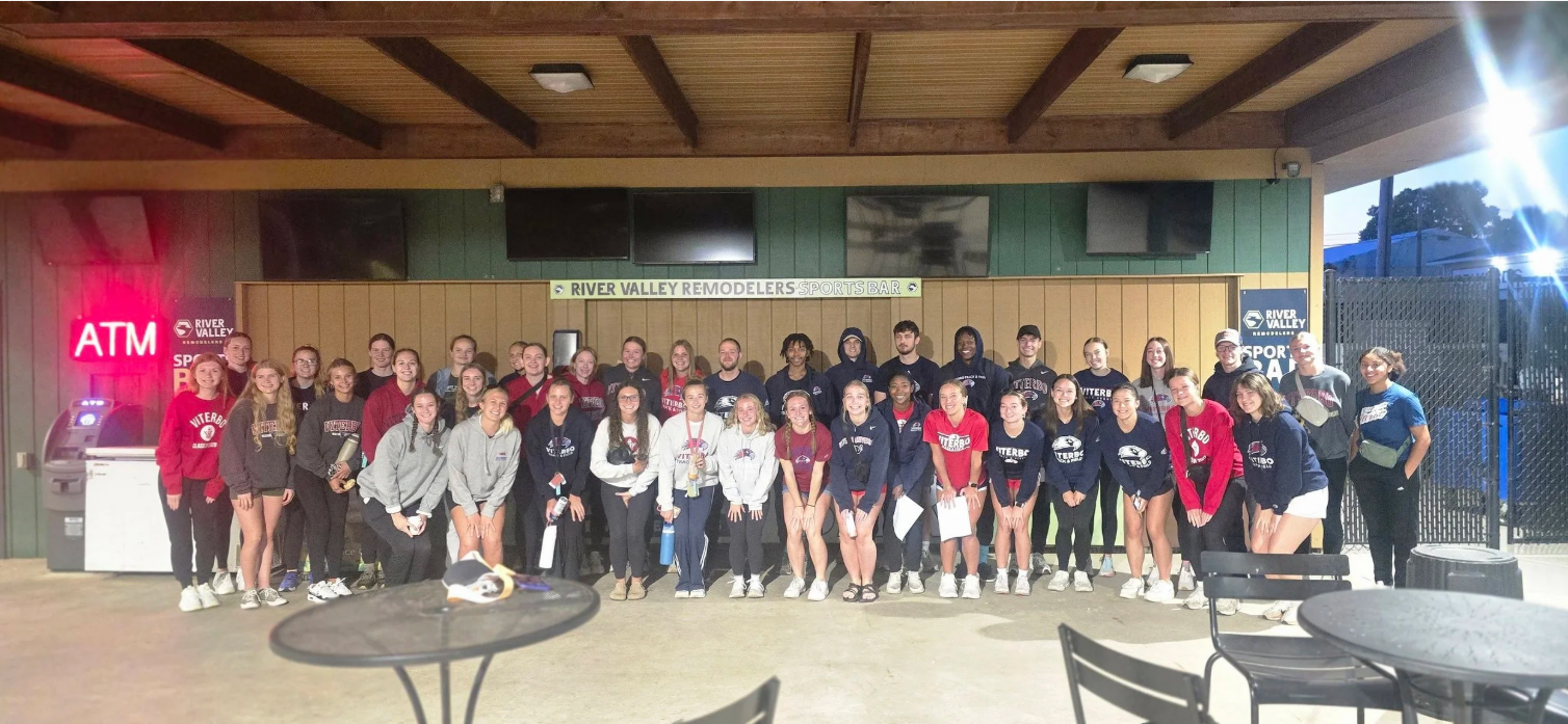 Group of young people posing for a photo in front of a sports bar with a sign that says 'River Valley Remodelers Sports Bar' in the background.