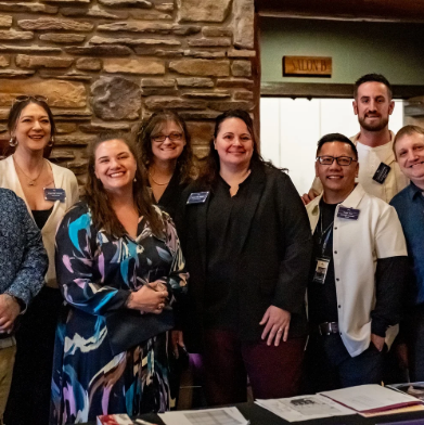 Group of seven smiling people at a restaurant or café, some wearing name tags, standing behind a table.