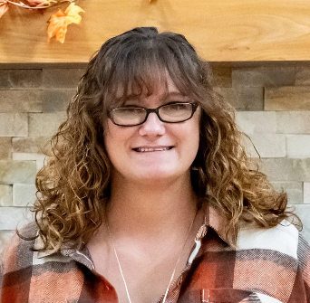 Woman with curly hair and glasses smiling, wearing a plaid shirt in front of a stone wall.