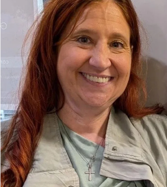 A woman with long red hair smiling, wearing a light gray jacket and a silver cross necklace.
