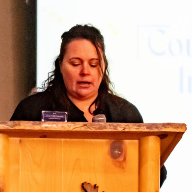 Woman standing at a wooden podium speaking into a microphone