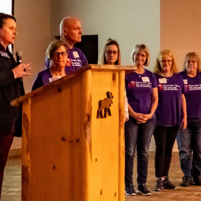 Group of six women standing next to each other in purple shirts, with one woman speaking into a microphone near a wooden podium in an indoor setting.