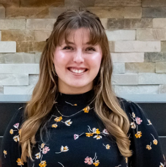 Young woman with long light brown hair and bangs, smiling, wearing a dark floral top, standing in front of a stone wall.