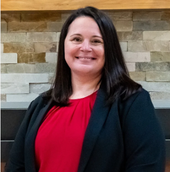 A woman with shoulder-length dark hair, wearing a red top and black blazer, smiling in front of a stone wall background.