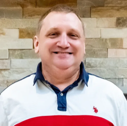 A smiling man wearing a white shirt with a red stripe, standing outdoors in front of a stone wall.