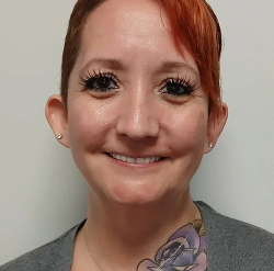 Close-up of a smiling woman with short red hair, wearing a gray top, standing against a plain background.