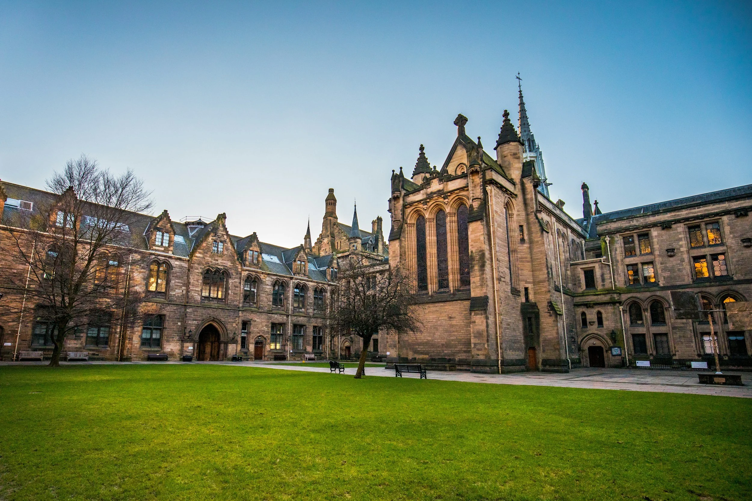 A historic stone building with gothic architecture and pointed spires, surrounded by a green lawn with benches and trees, under a blue sky.