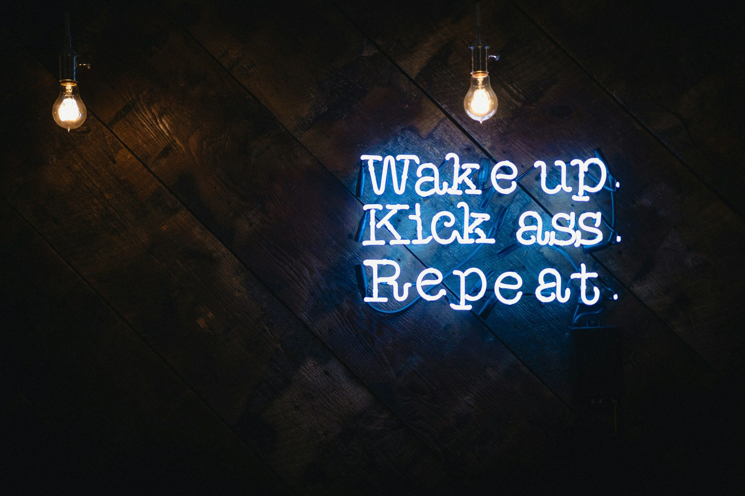 Neon sign on dark wood wall reads 'Wake up. Kick ass. Repeat.' with two lightbulbs hanging above.