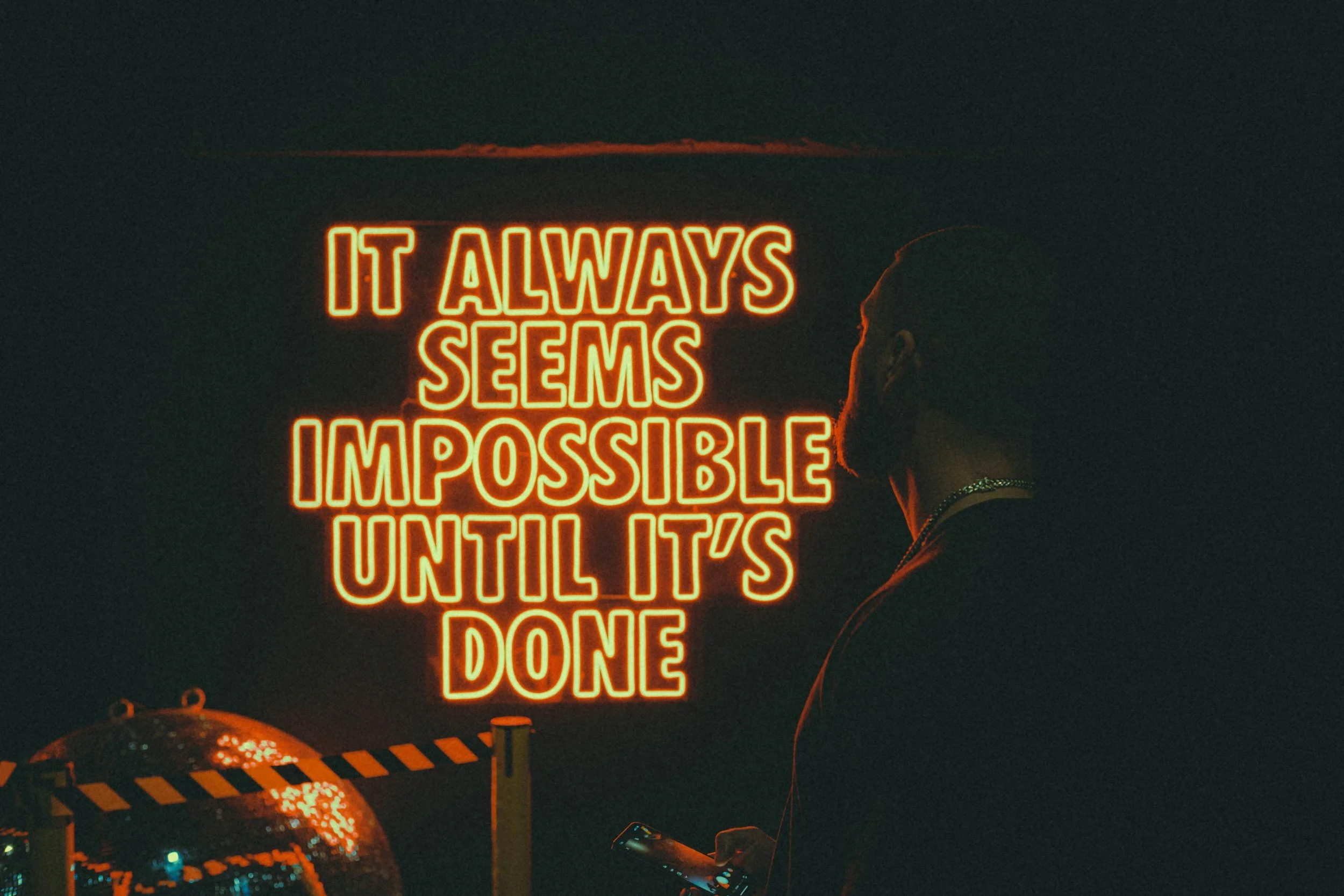 A neon sign in orange that reads, "It always seems impossible until it's done," with a person standing in front of it, looking at their phone in a dark environment.