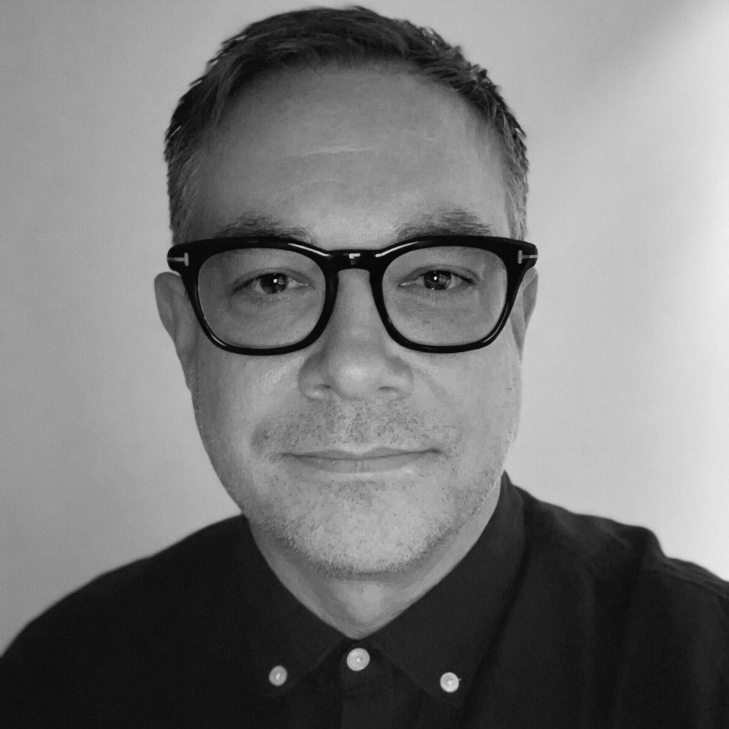 Portrait of a man with glasses, short hair, and a slight smile, wearing a dark shirt with a button-down collar.