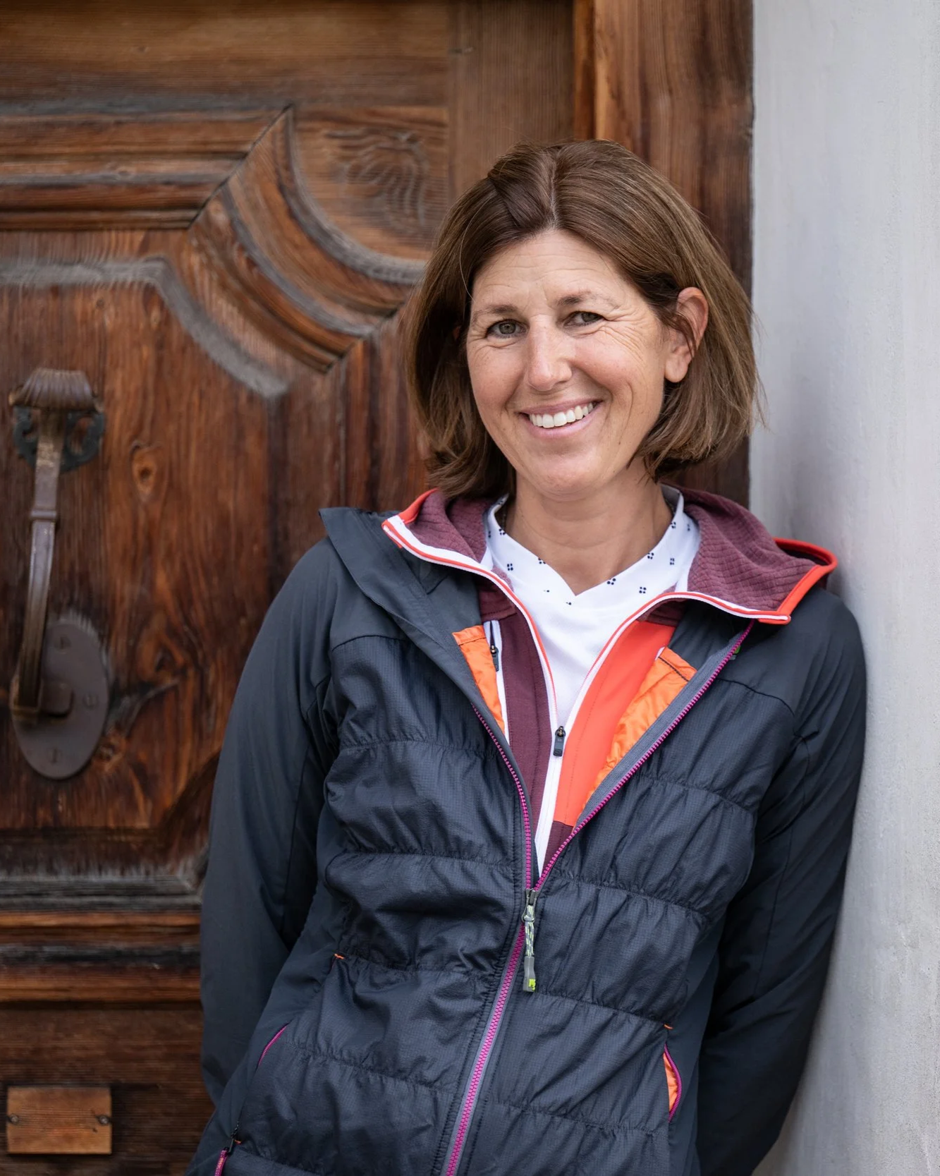 Eine lachende Frau steht neben einer Holztür, trägt eine dunkelblaue Jacke mit orange Details und ein weißes T-Shirt.