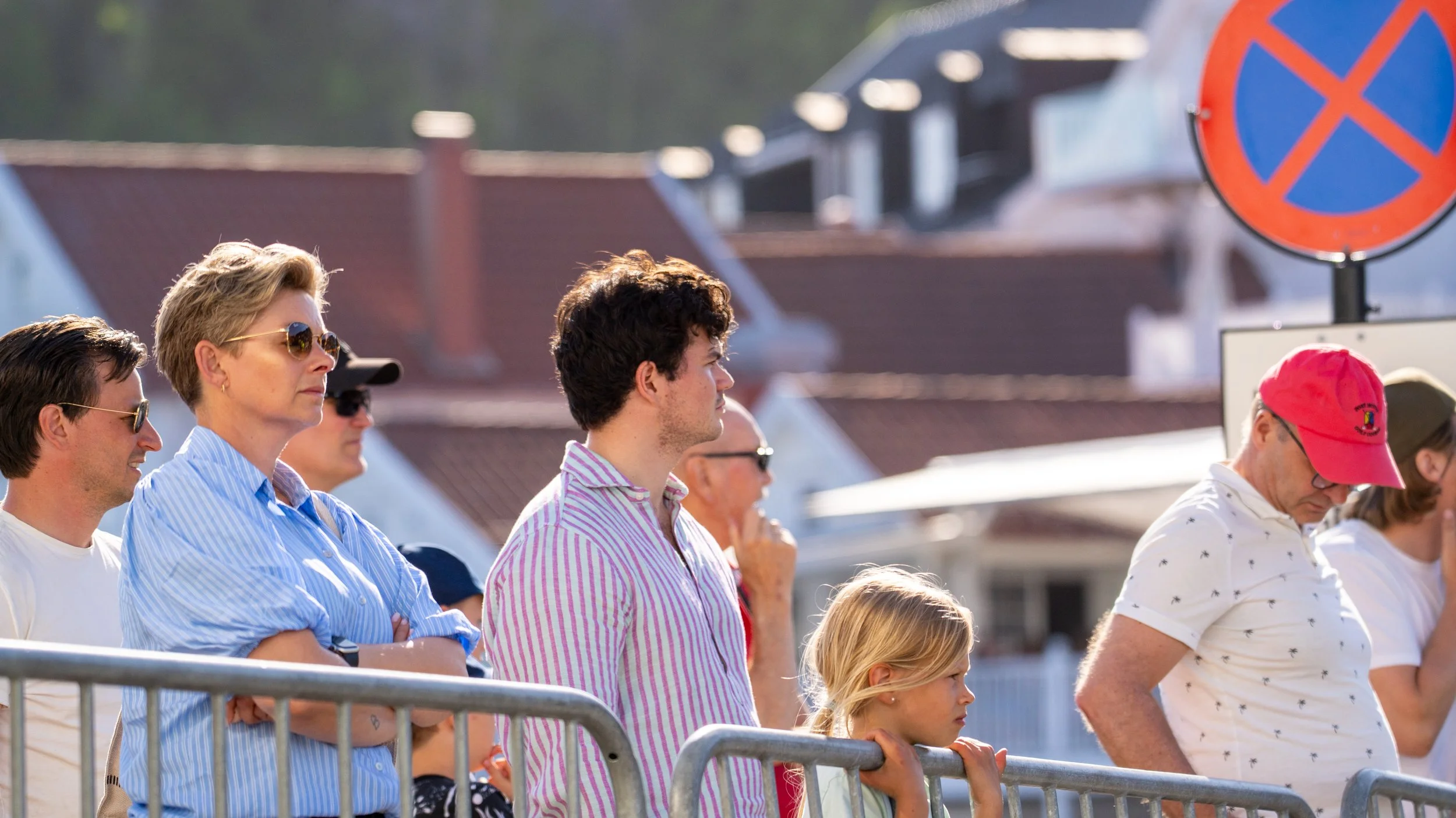 Group of people standing behind a metal barrier, some wearing sunglasses, in an outdoor setting with houses in the background.