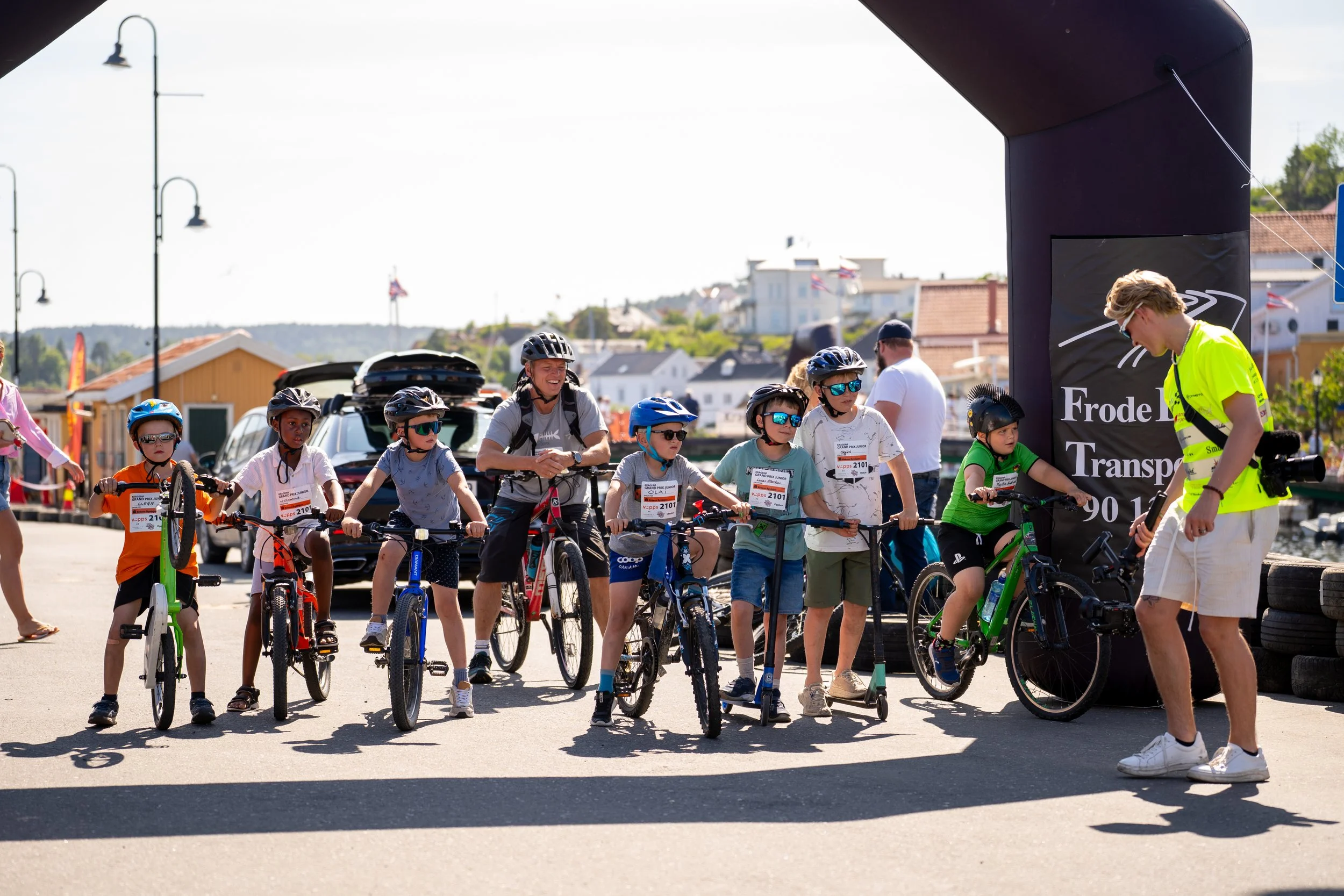 Children with bikes and helmets gathered at the starting line of a community bike race, under a black inflatable arch with event signage, on a sunny day.
