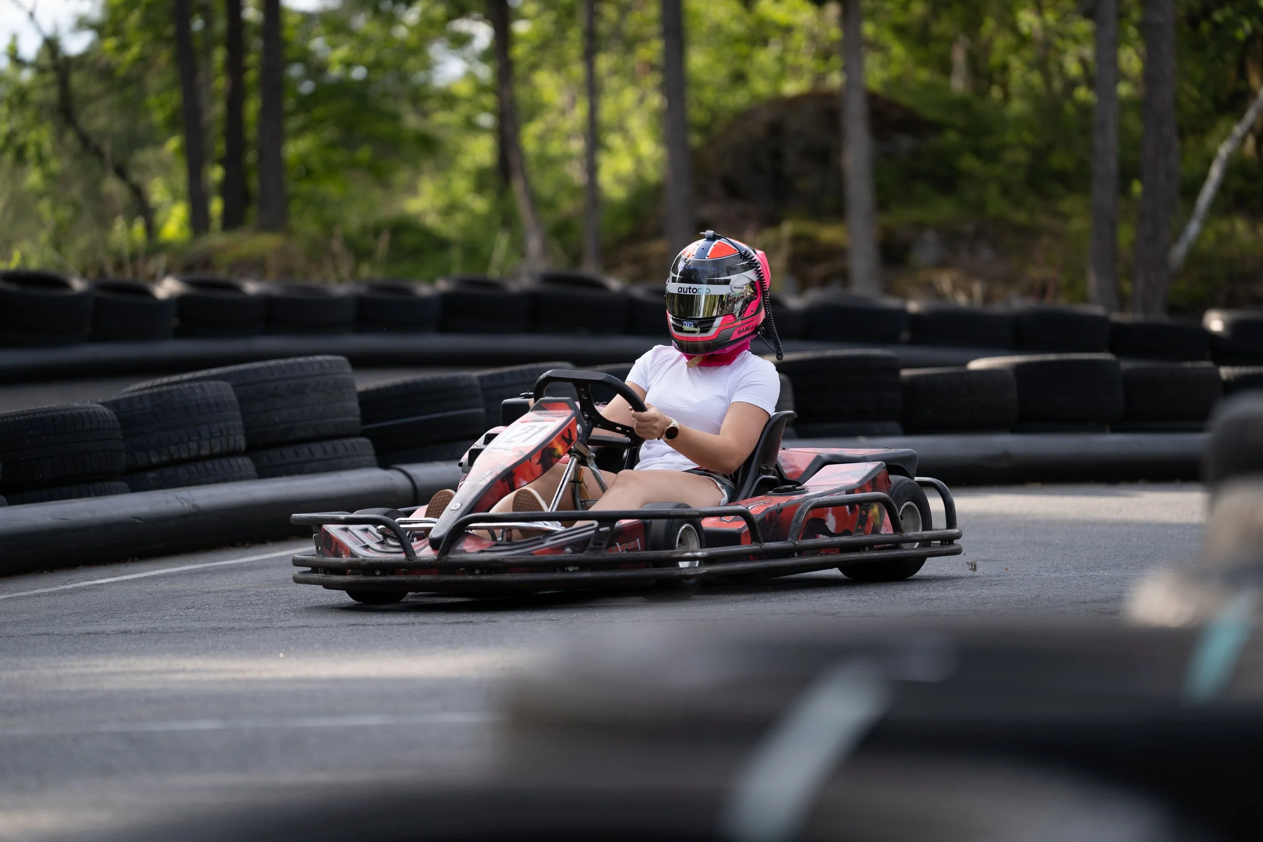 A person wearing a helmet and white shirt is driving a go-kart on a race track surrounded by tire barriers, with a wooded area in the background.