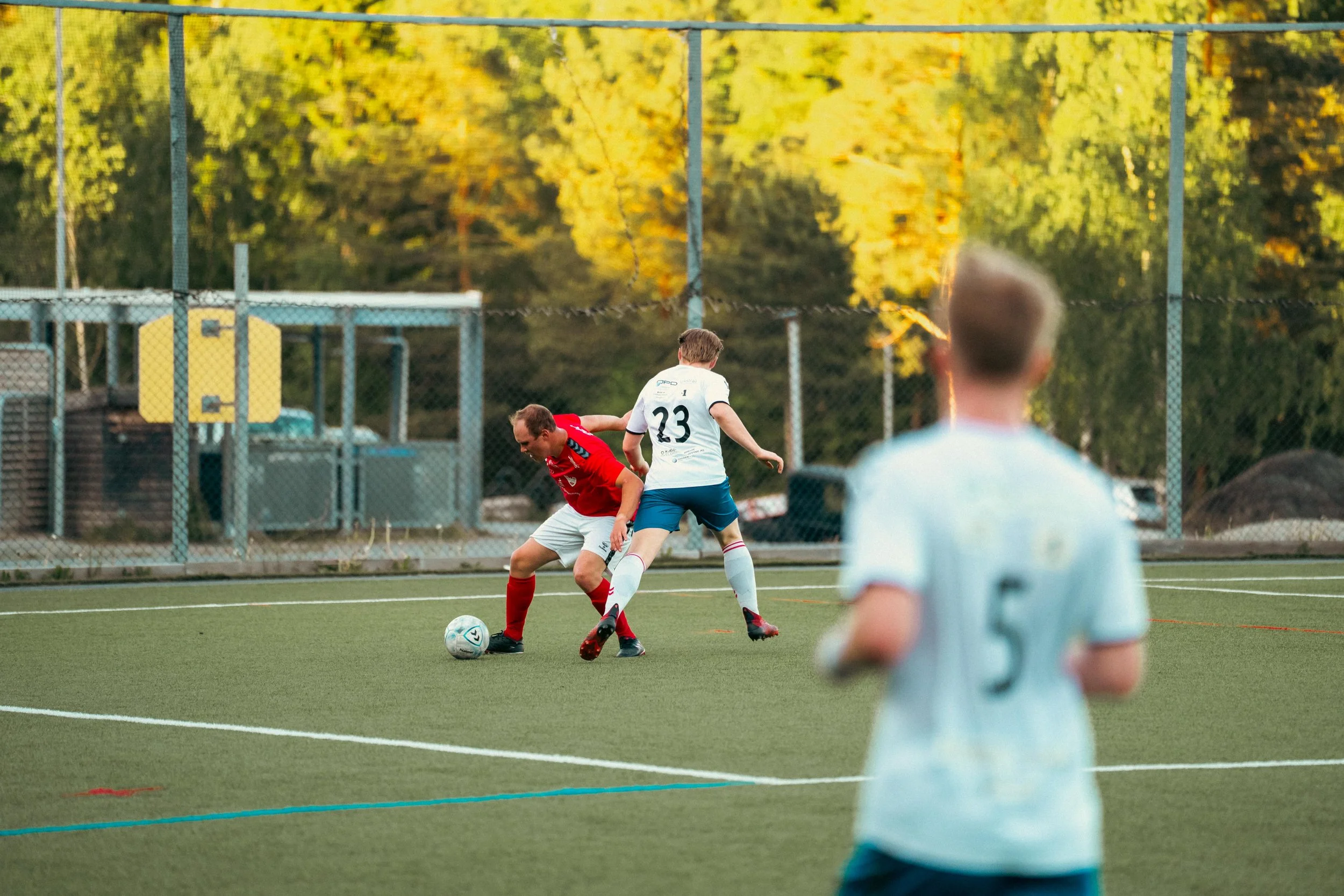 Two young boys playing soccer on a green field, one in a red jersey and white shorts, the other in a white jersey with the number 23, contesting for the ball. A third player in a light blue jersey with the number 5 is in the foreground, blurred.