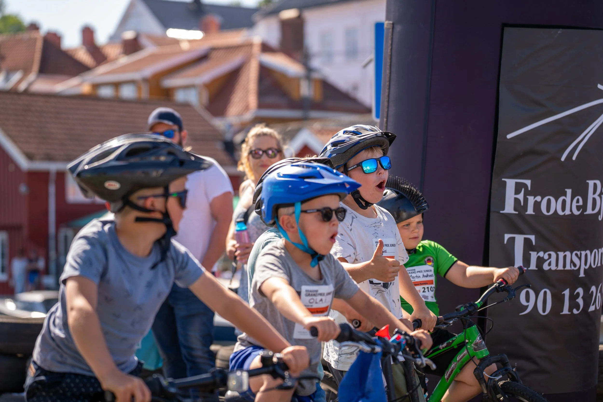 Group of children in helmets and sunglasses riding bikes at a race event, with adults and houses in the background.