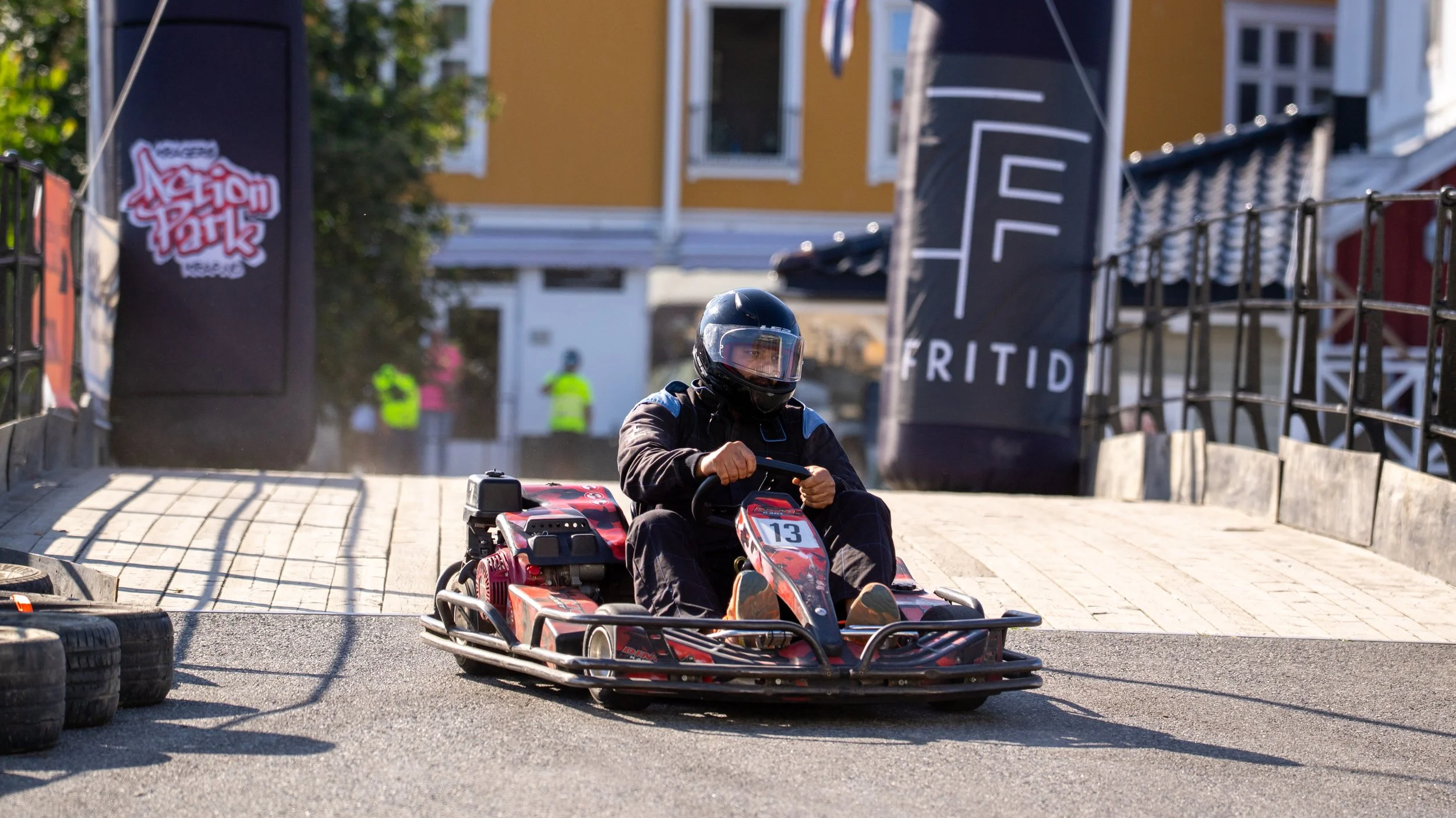 Person driving a go-kart on an outdoor track, wearing a black helmet and protective gear, with some tires and banners visible on the side.