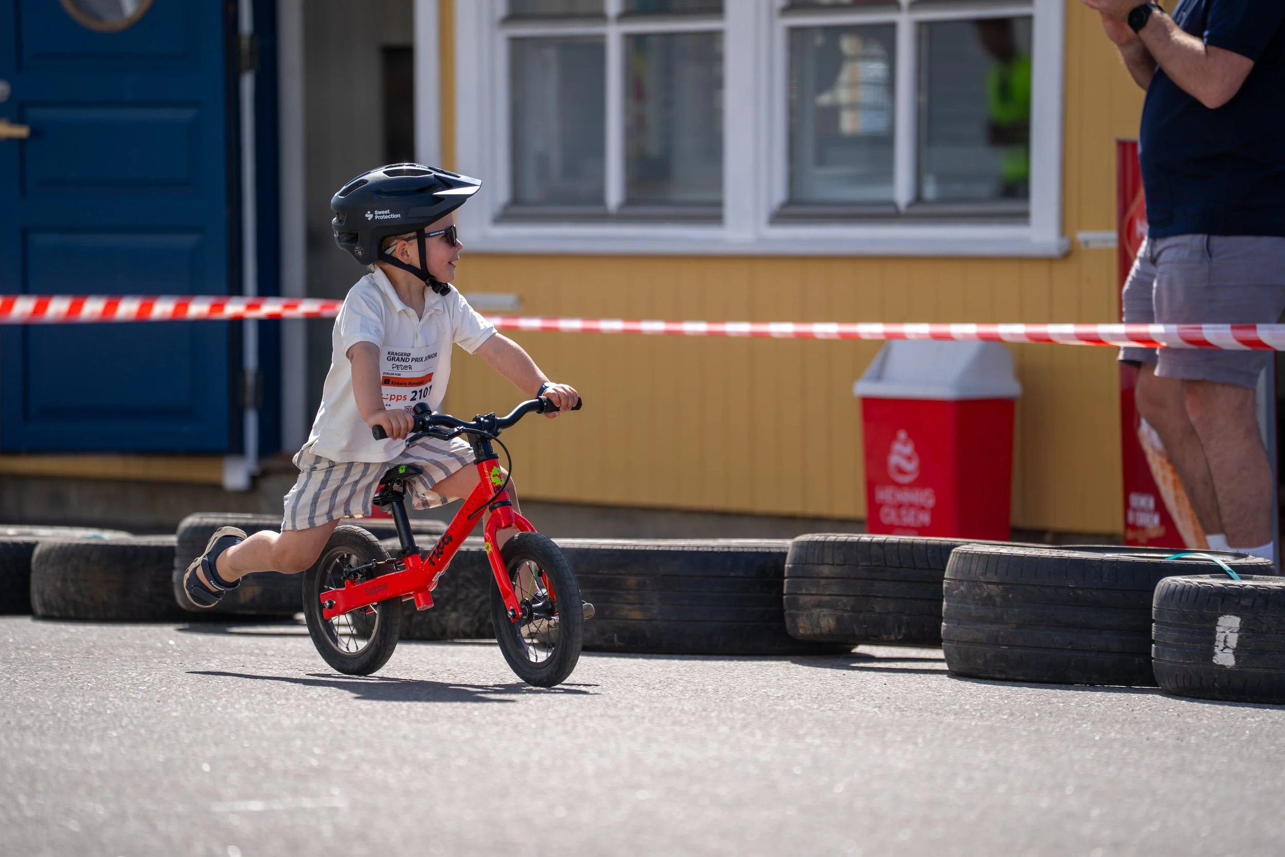 A young boy wearing a helmet riding a red balance bike on a paved surface during a race or event, with tires and a barrier in the background.