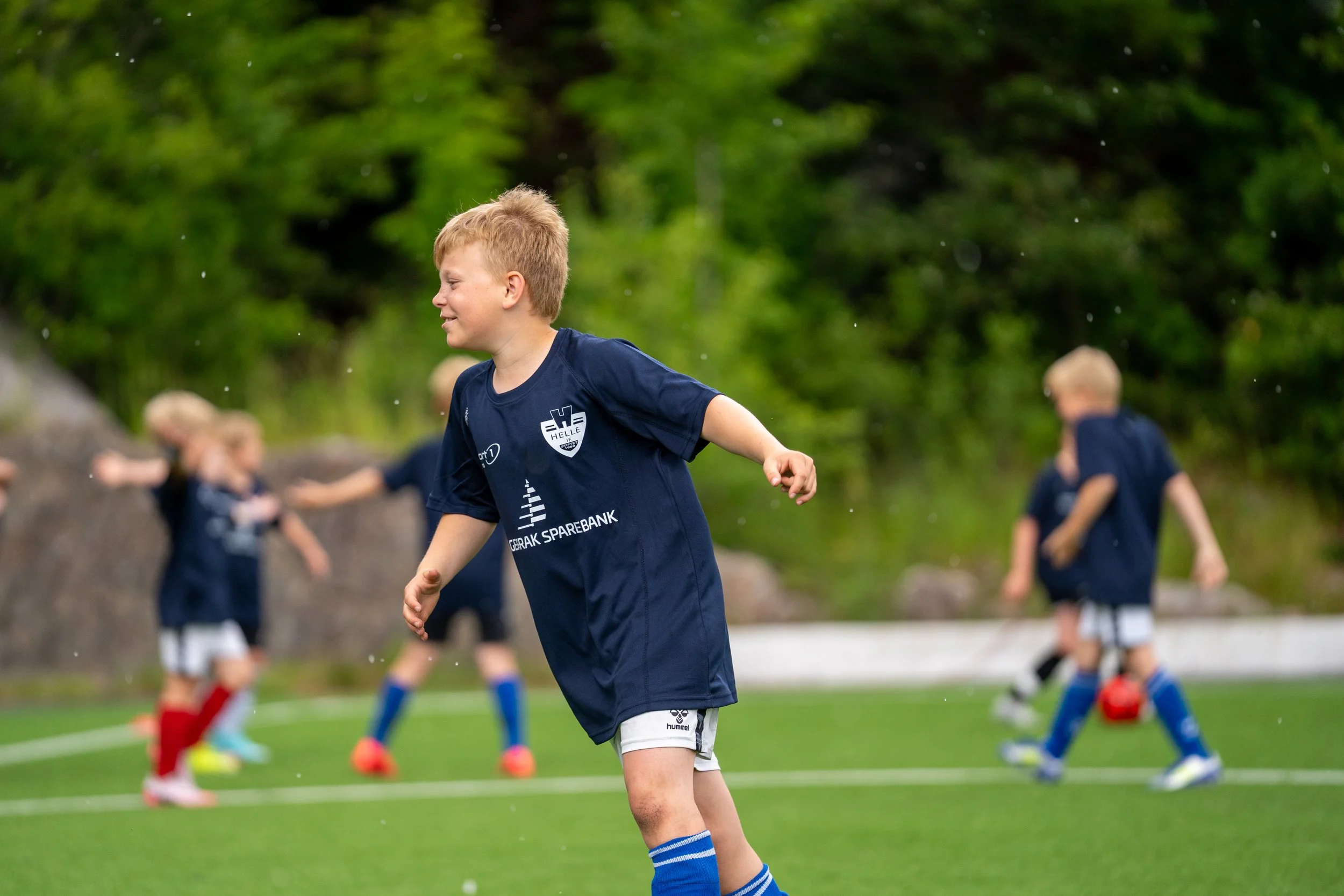 Young boys playing soccer on a green field, wearing navy blue jerseys and shorts, with trees in the background.