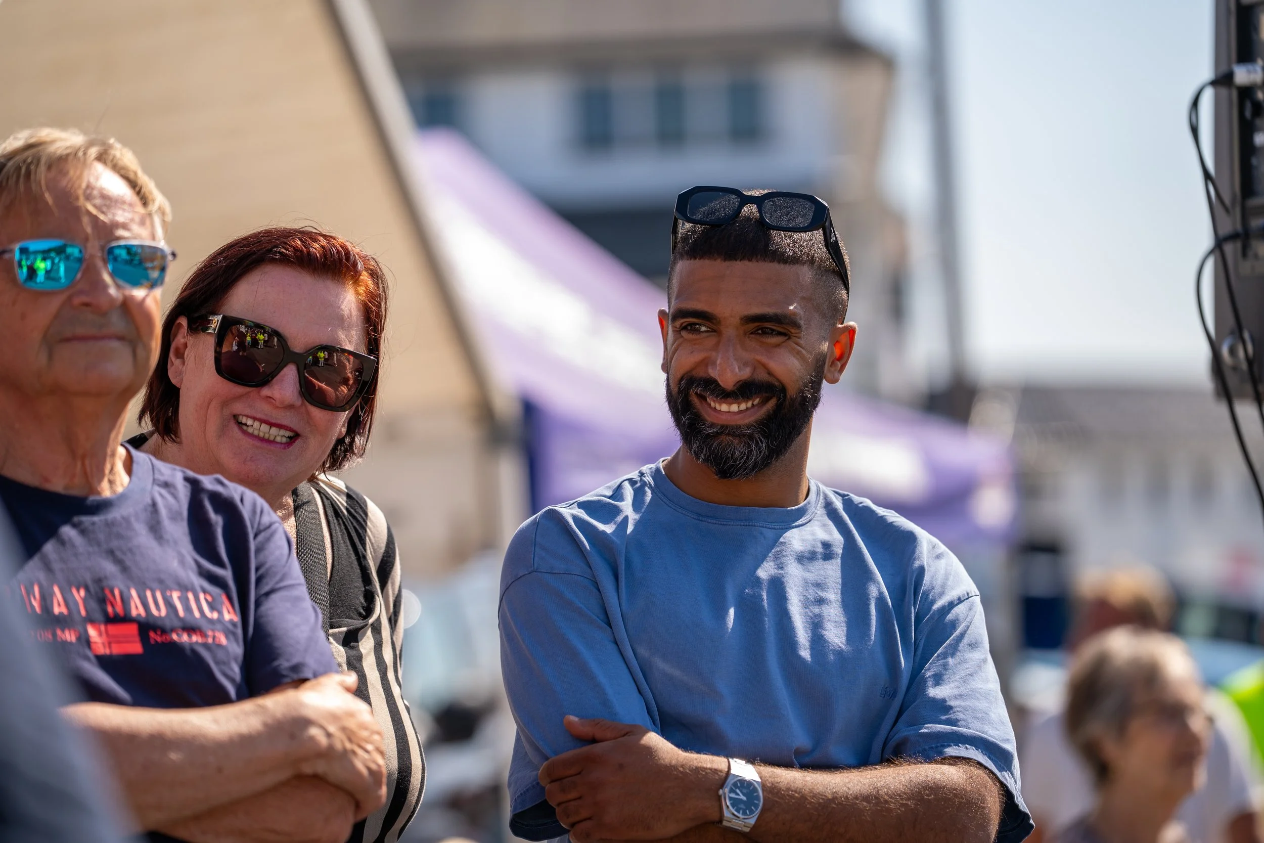 Three people smiling outdoors during daytime, two women and one man, with boats and a waterfront in the background.