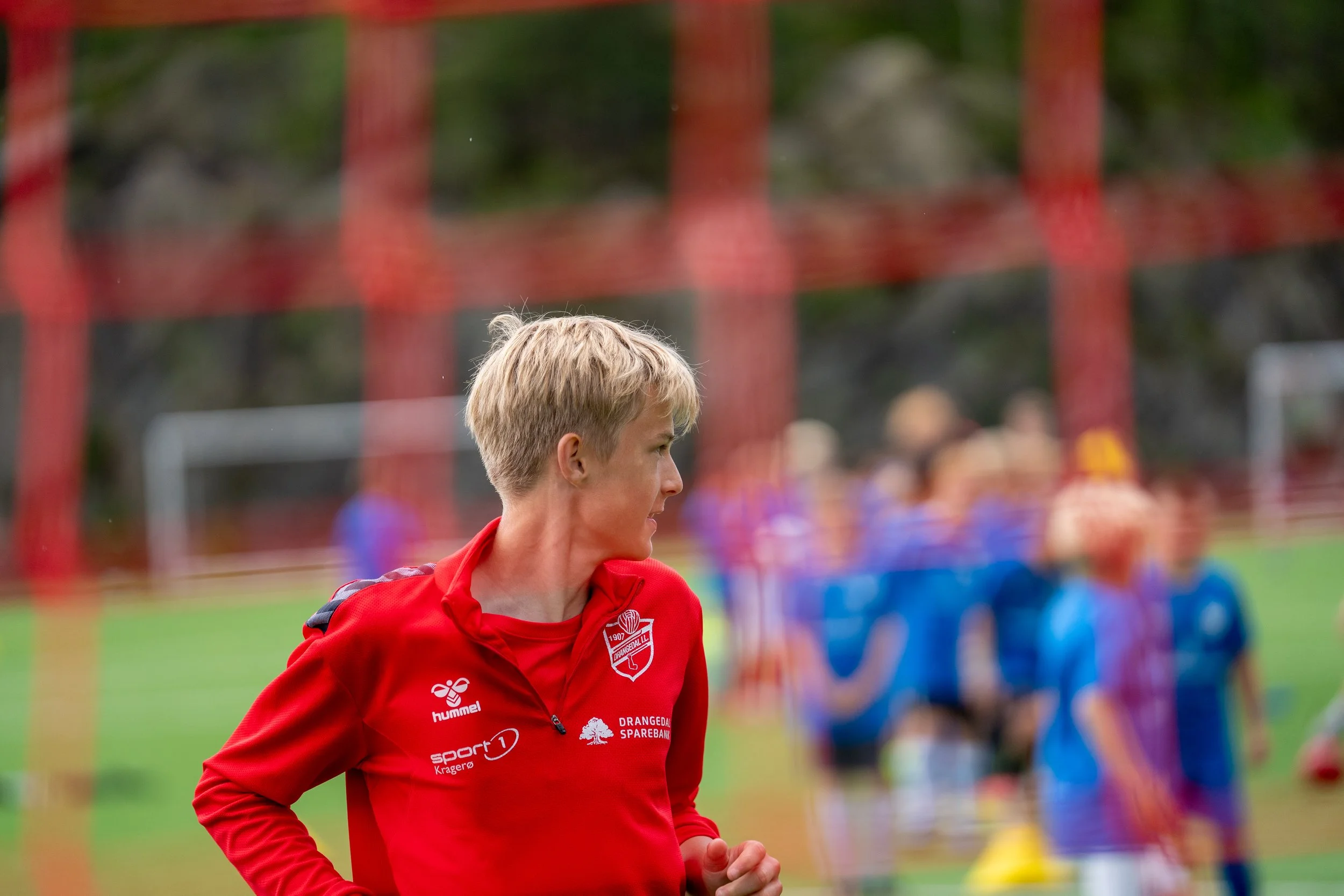 A female soccer coach or trainer in a red jacket on the field with a group of children in sports uniforms in the background.