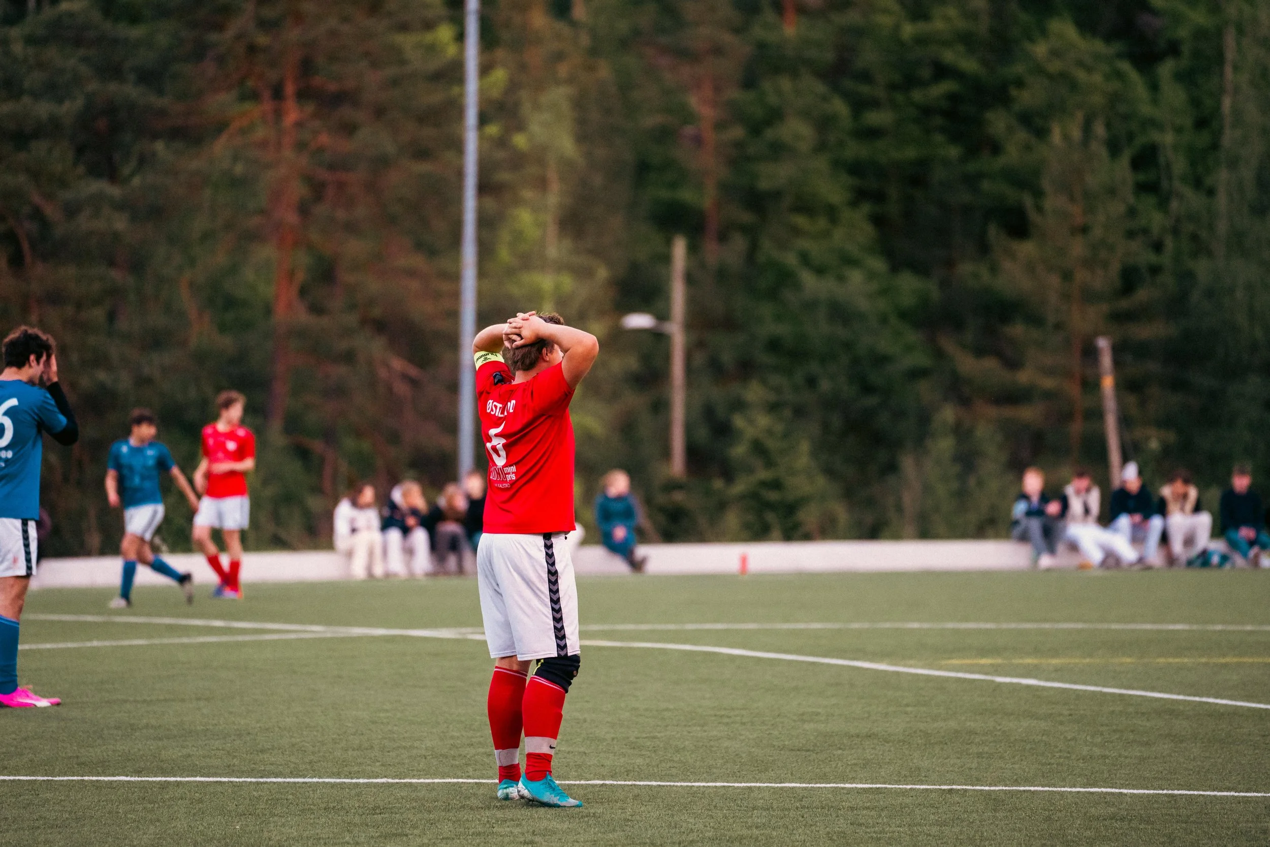 A young soccer player in a red jersey and red socks stands with hands on his head on a soccer field during a game, with other players and spectators in the background.
