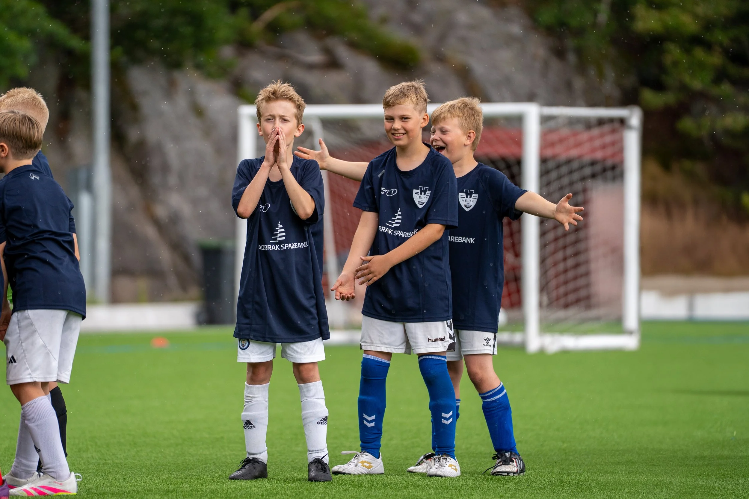 Group of young boys in navy blue soccer jerseys celebrating on a soccer field with a goalpost in the background.