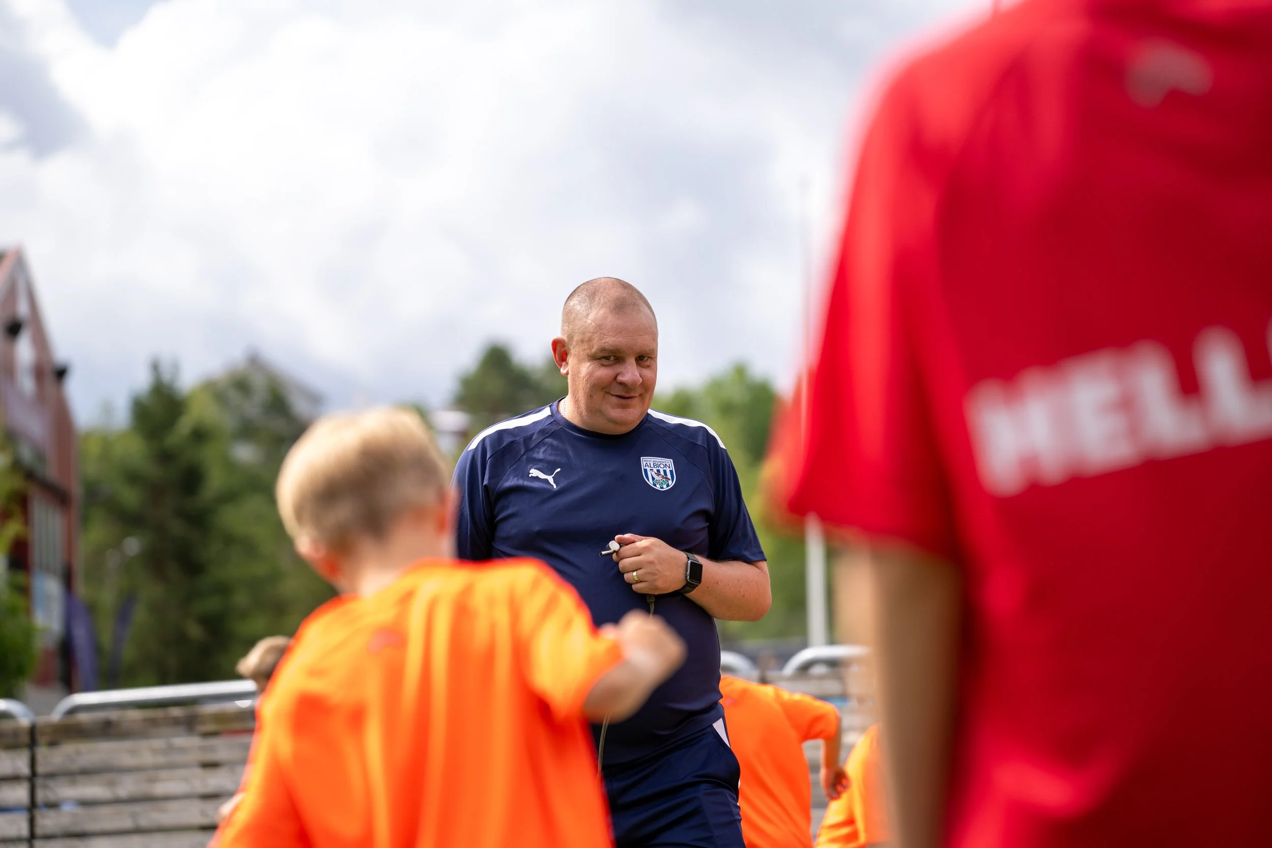 A man in a navy blue athletic shirt with the Albion football club logo and a Puma logo stands outdoors, surrounded by children in orange sports jerseys. The man is smiling slightly and holding a whistle, with trees and a cloudy sky in the background.