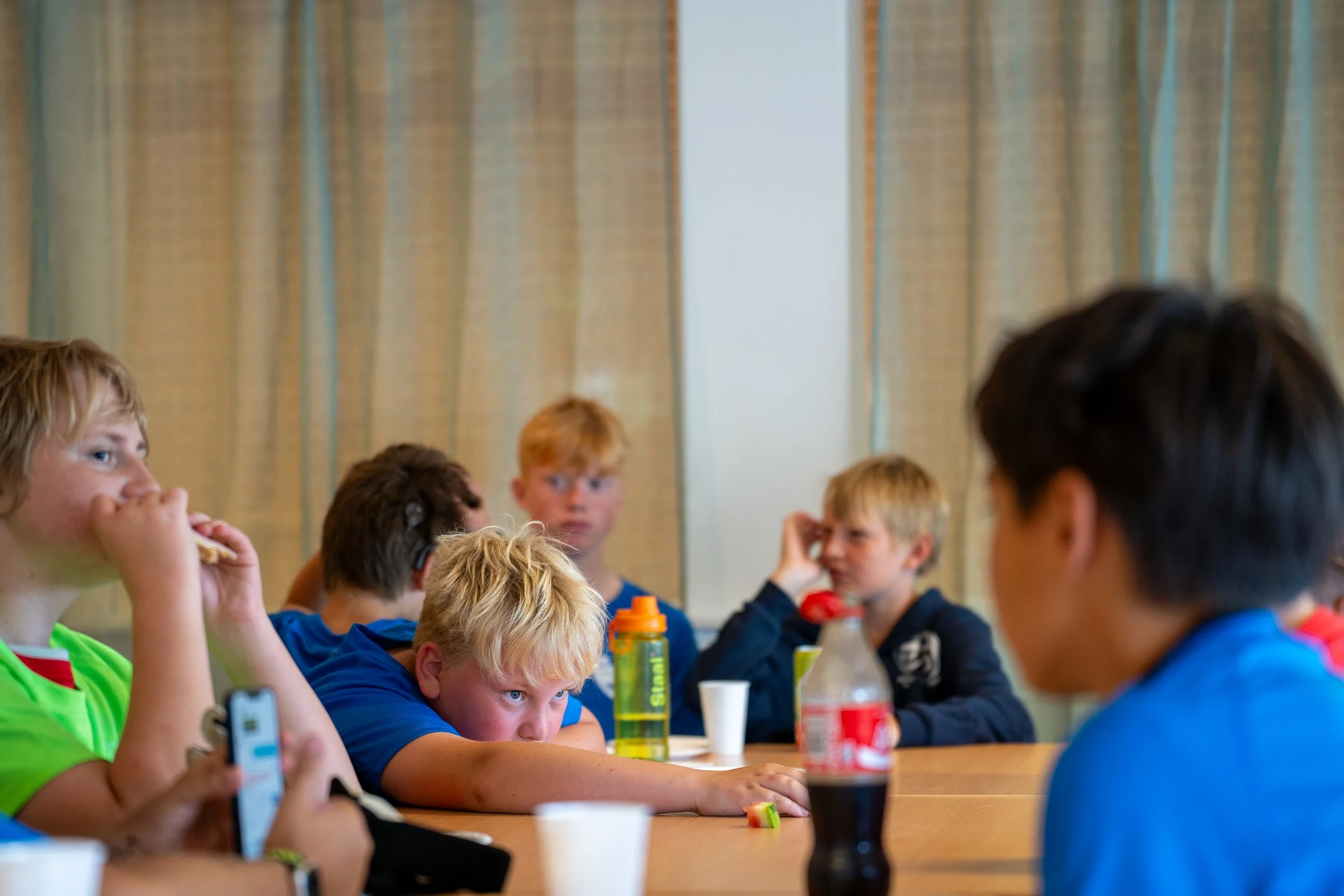 Group of children sitting at a table in a room with beige curtains, some are drinking, some appear bored or distracted.