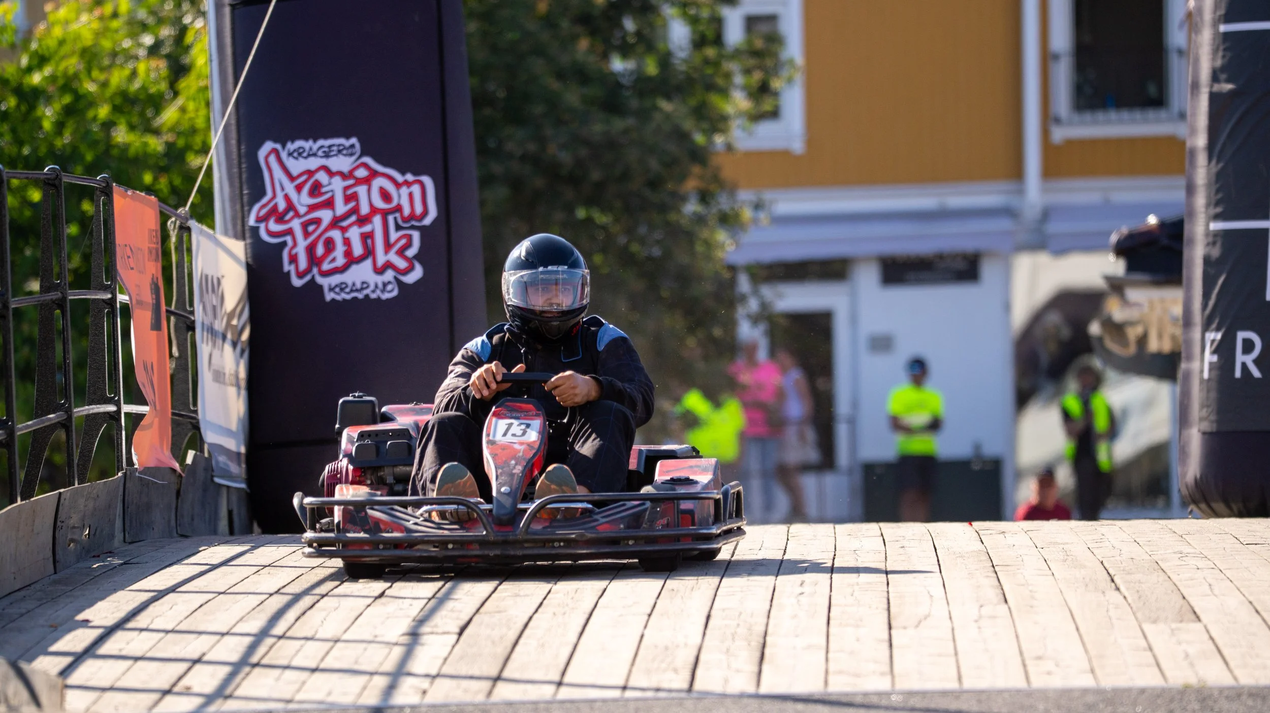 A person wearing a helmet and racing suit driving a go-kart on a track at Action Park, with spectators and staff in the background.