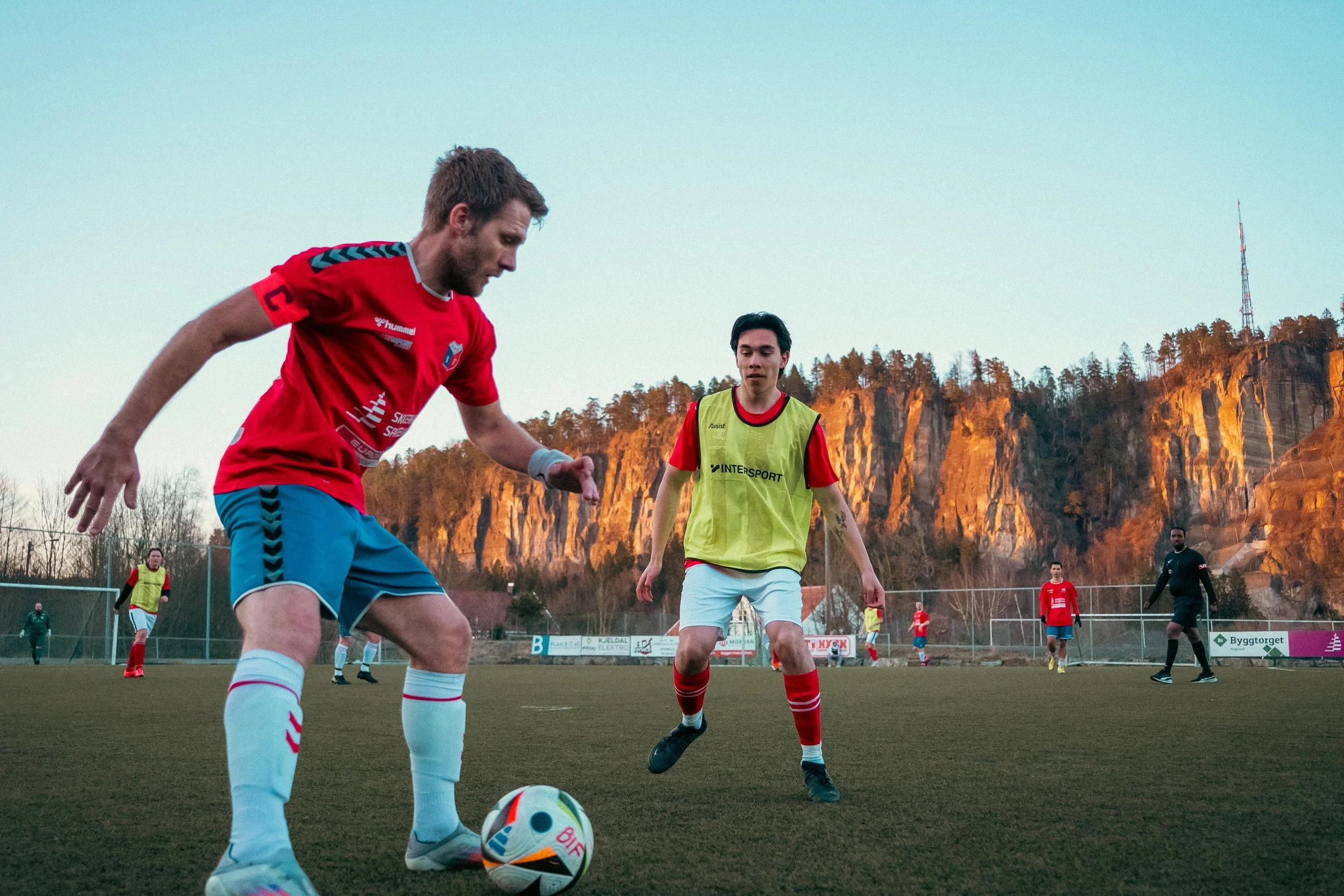 Two soccer players, one in a red jersey and blue shorts, and the other in a yellow bib and white shorts, on a soccer field during sunset with rocky cliffs and trees in the background.