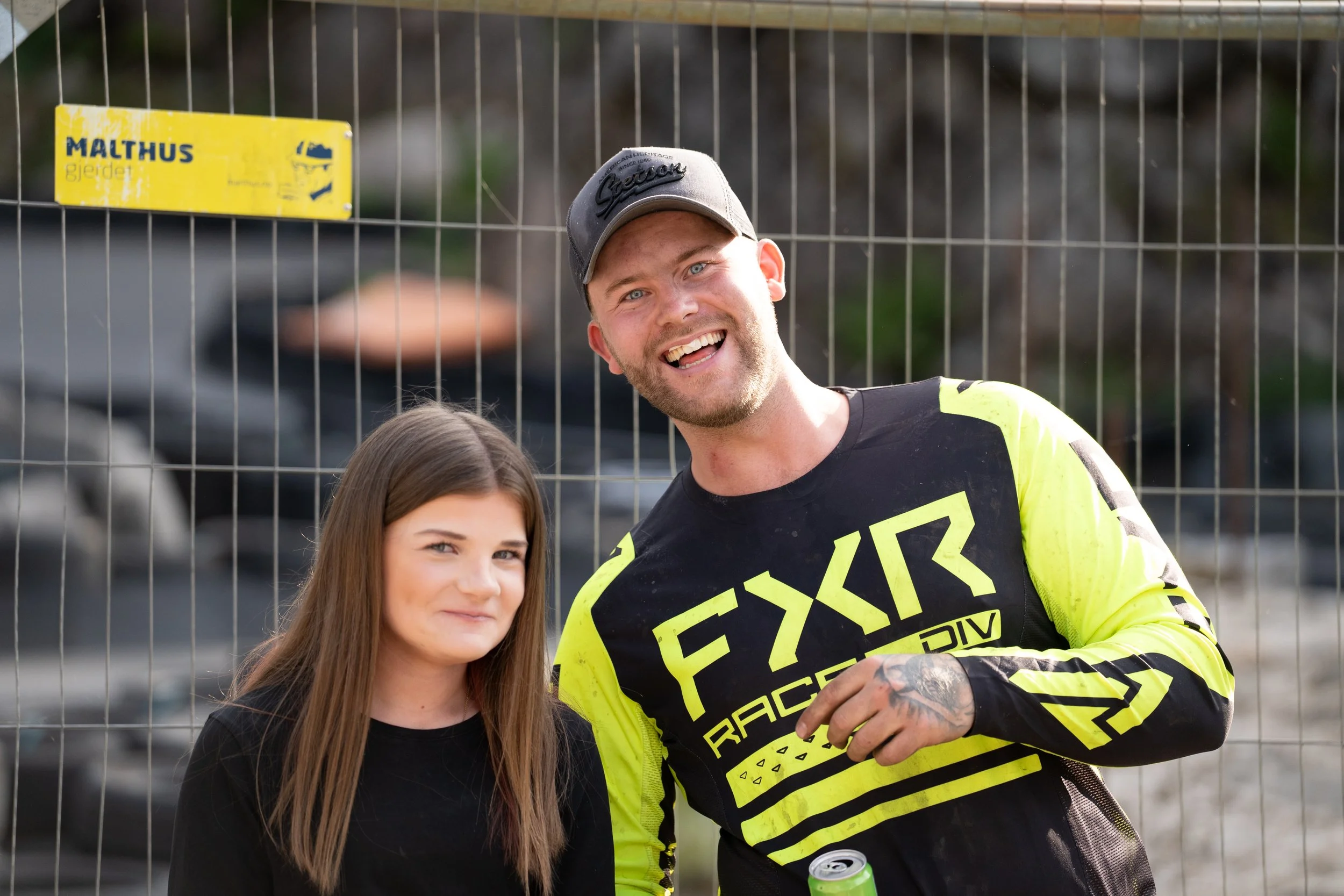 A young woman with long brown hair and a man with a beard wearing a gray cap and a bright yellow and black FXR Racing dirt bike jersey standing in front of a wire fence, smiling for the camera, with a blurred outdoor background.