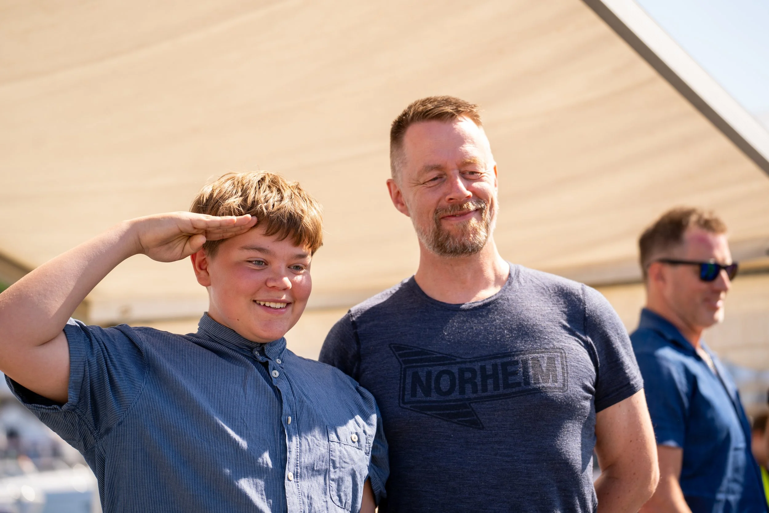 A young boy saluting next to a man in a gray shirt with a logo, outdoors with a sunny sky.