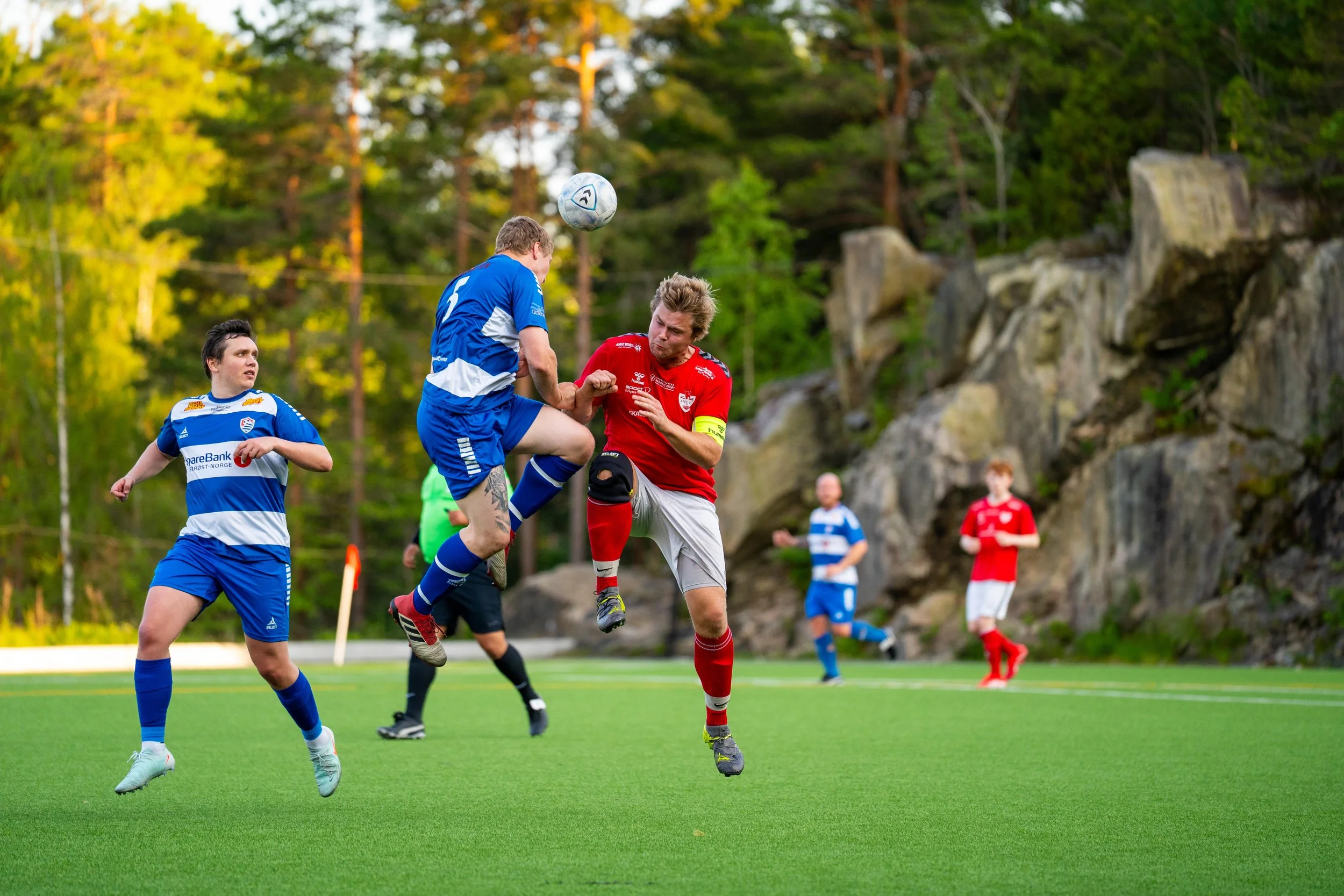 Soccer players in blue and red uniforms competing for the ball during a match on a green field, with trees and rocks in the background.