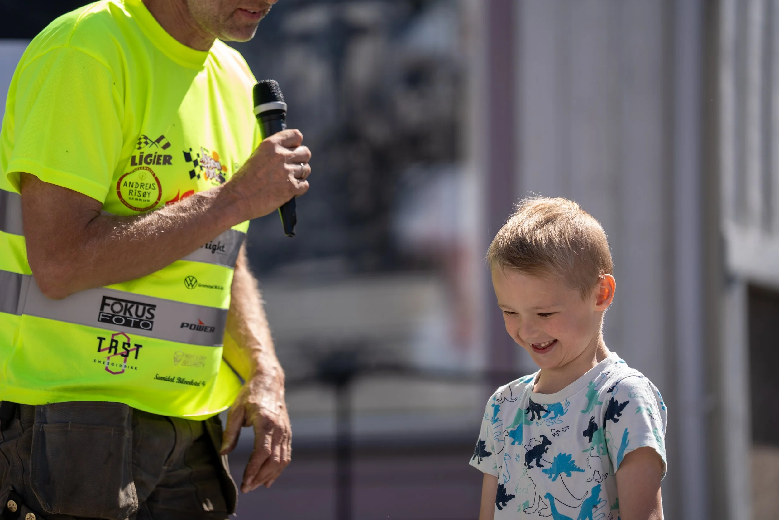 A man in a bright yellow t-shirt holding a microphone and talking to a young boy who is smiling and looking down, outdoors on a sunny day.