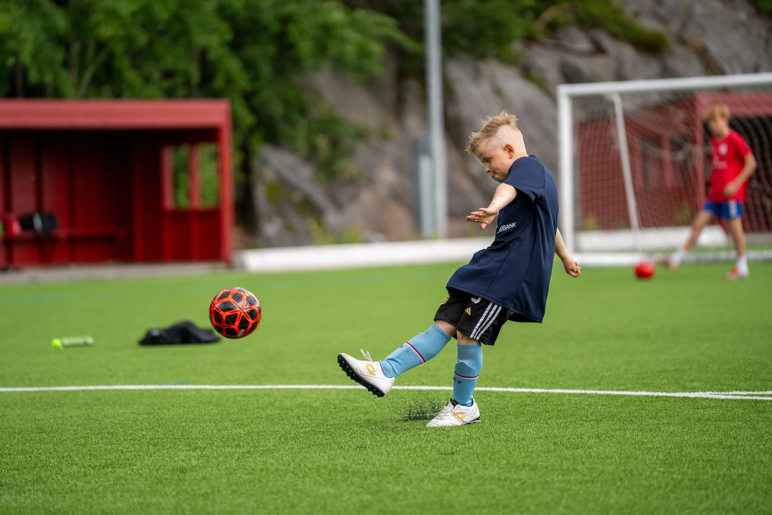 A young boy wearing a blue sports uniform and soccer socks kicks a black and red soccer ball on a green field. In the background, another child in a red shirt and shorts stands near a goalpost.