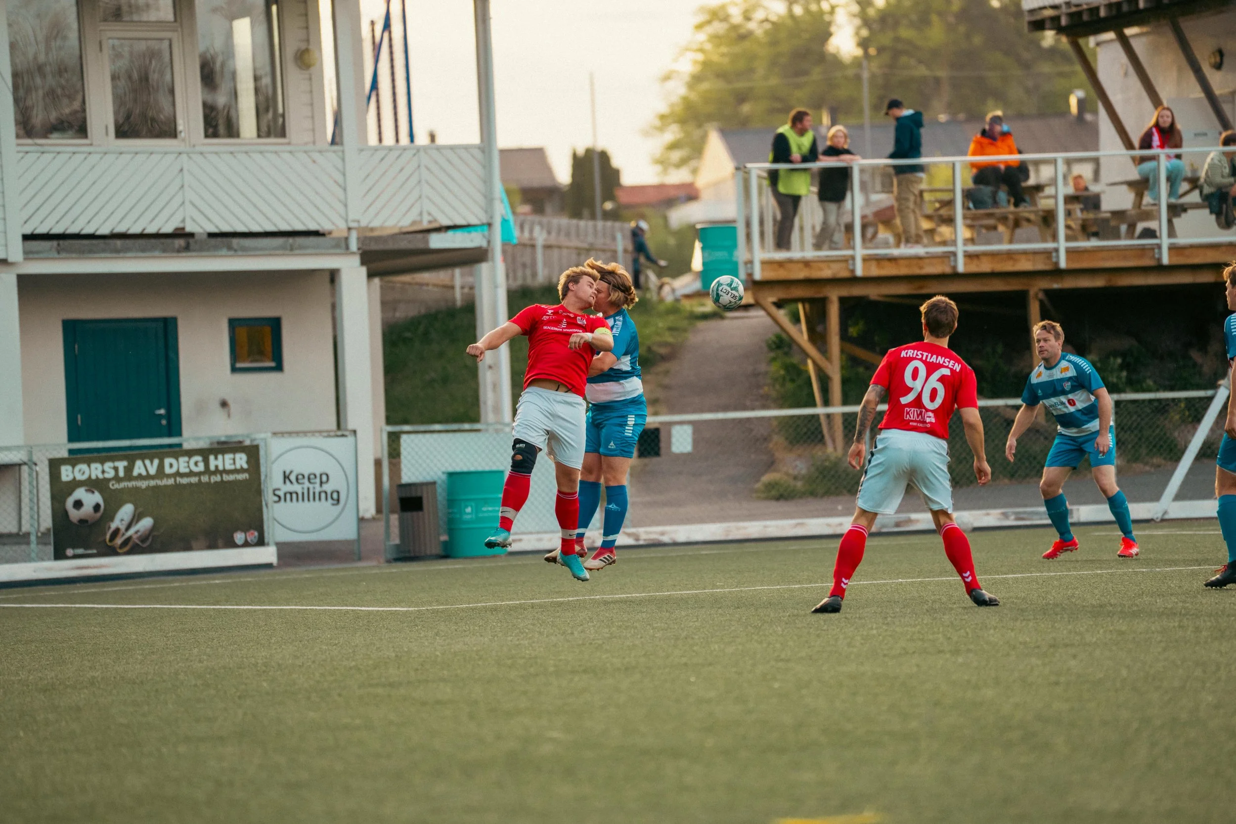 Two soccer players in mid-air contest for the ball, with others watching on the field and spectators in the stands during a game.