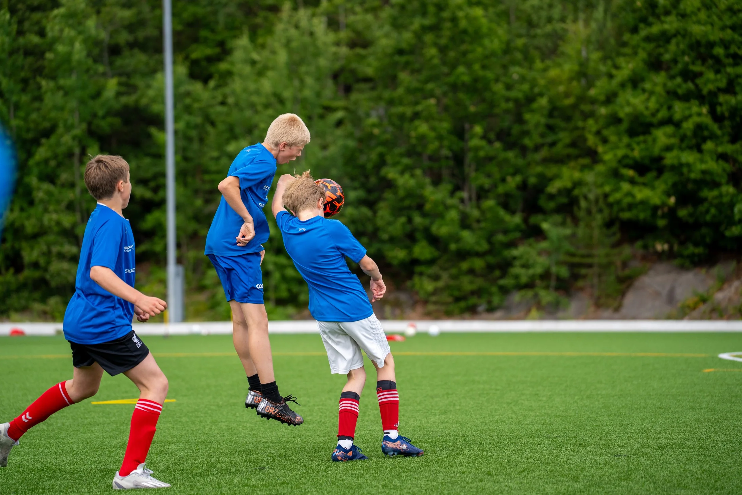 Four boys playing soccer on a green field, with trees in the background, wearing blue shirts and athletic shoes.