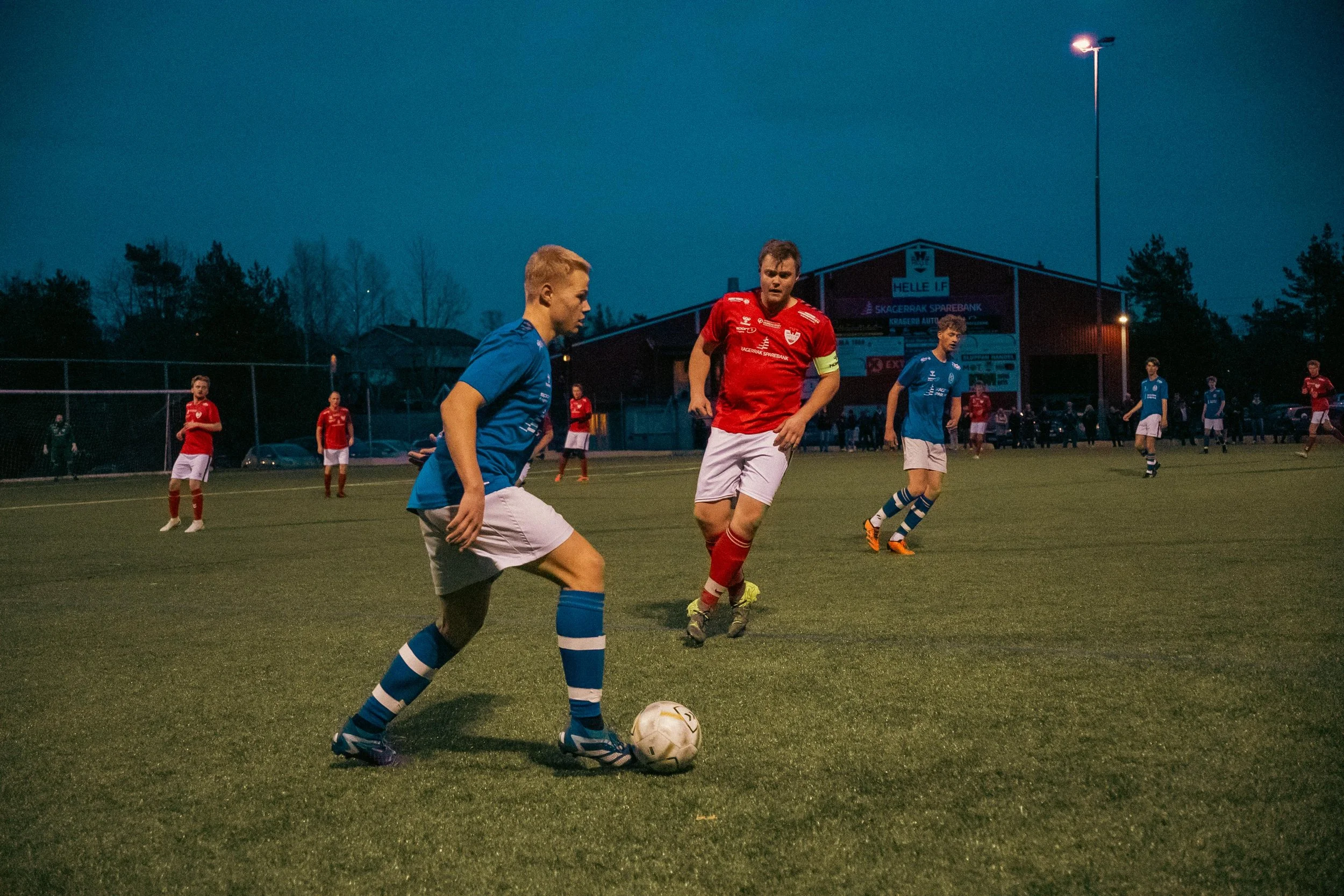 Soccer players on the field during a match at dusk, with one player in blue controlling the ball and others in red and blue in the background.