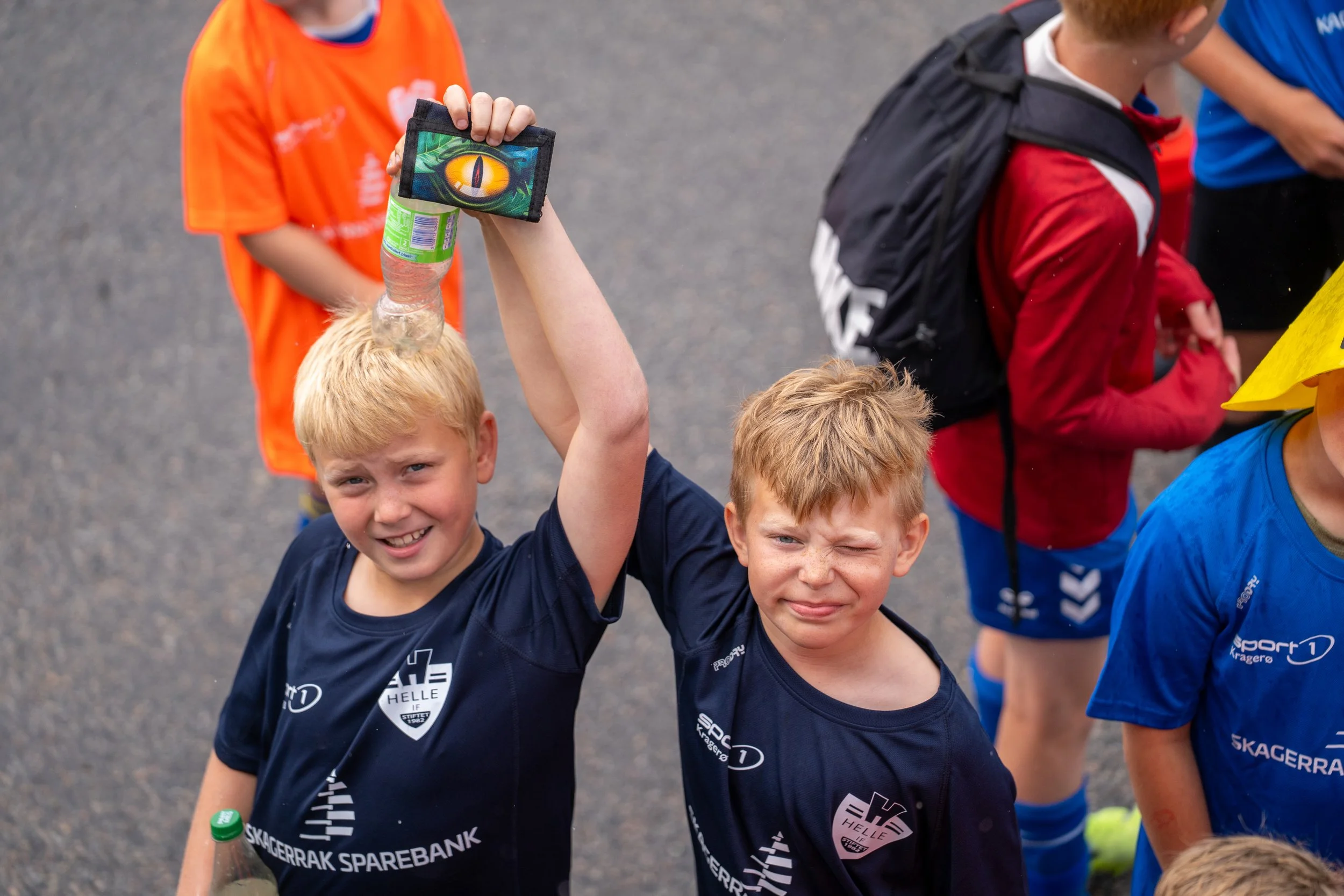 Two young boys with blonde hair, wearing dark blue sports jerseys with the words 'Skagerak Sparebank,' stand close together. The boy on the left is smiling, holding a water bottle, and raising his arm. The boy on the right is making a playful face. O