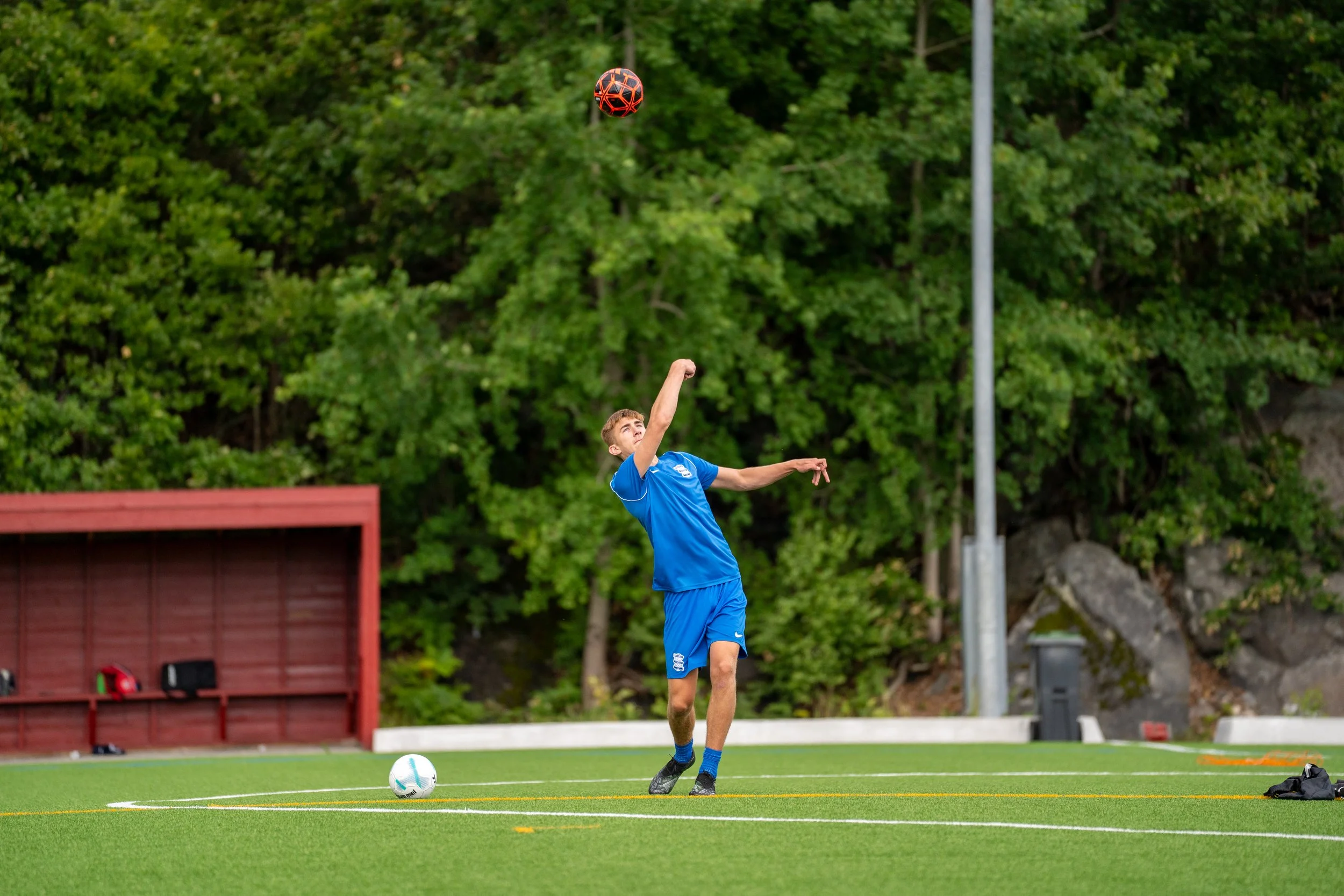 Young male soccer player in blue uniform kicking a soccer ball on a green field with trees in the background.