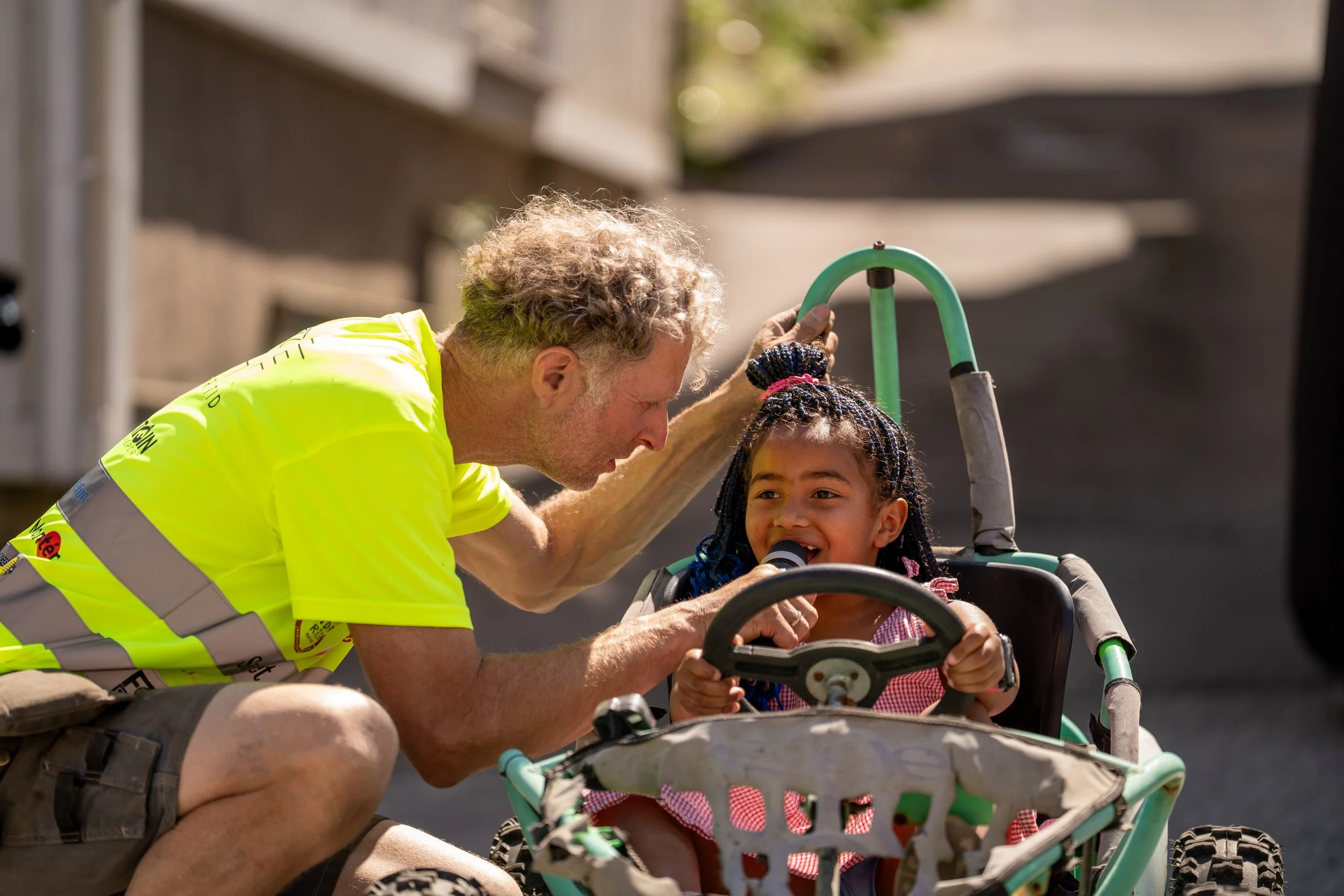 An older man and a young girl with braided hair are outside. The girl is sitting in a stroller and holding a toy guitar, smiling. The man is leaning over her, touching her head with his hand, and talking to her. The scene appears sunny and cheerful.