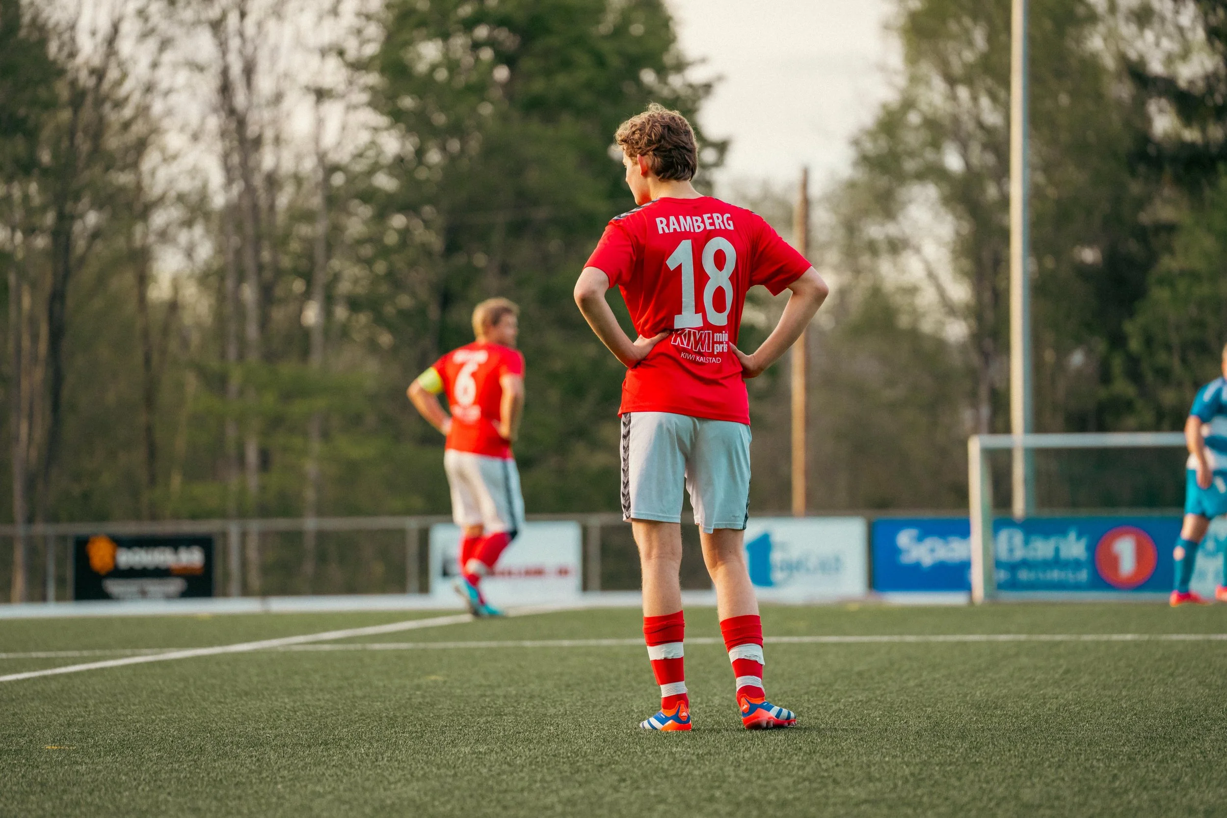 A soccer player wearing a red jersey with the name 'Ramberg' and the number 18 is standing on the field with hands on hips, looking towards the goal. Another player in a red jersey and a goalkeeper in a blue uniform are visible in the background, wit