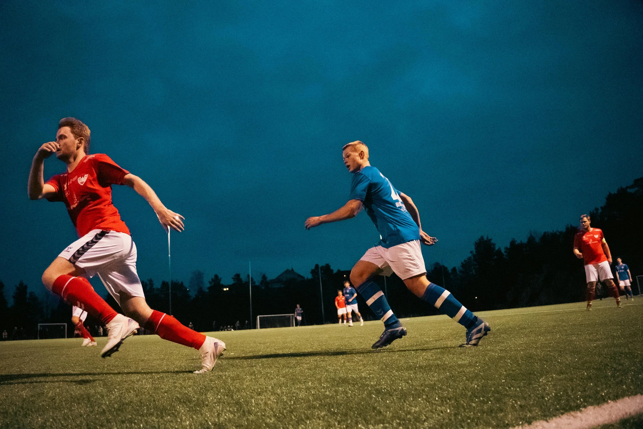 Soccer players in red and blue uniforms on a field during a night game.