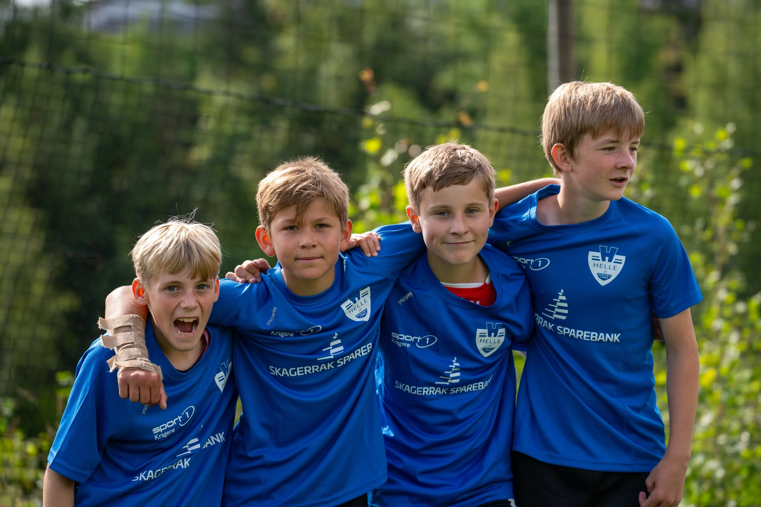 Four young boys in blue sports jerseys standing together outdoors, with their arms around each other's shoulders, smiling and posing for a photo on a grassy field with trees in the background.