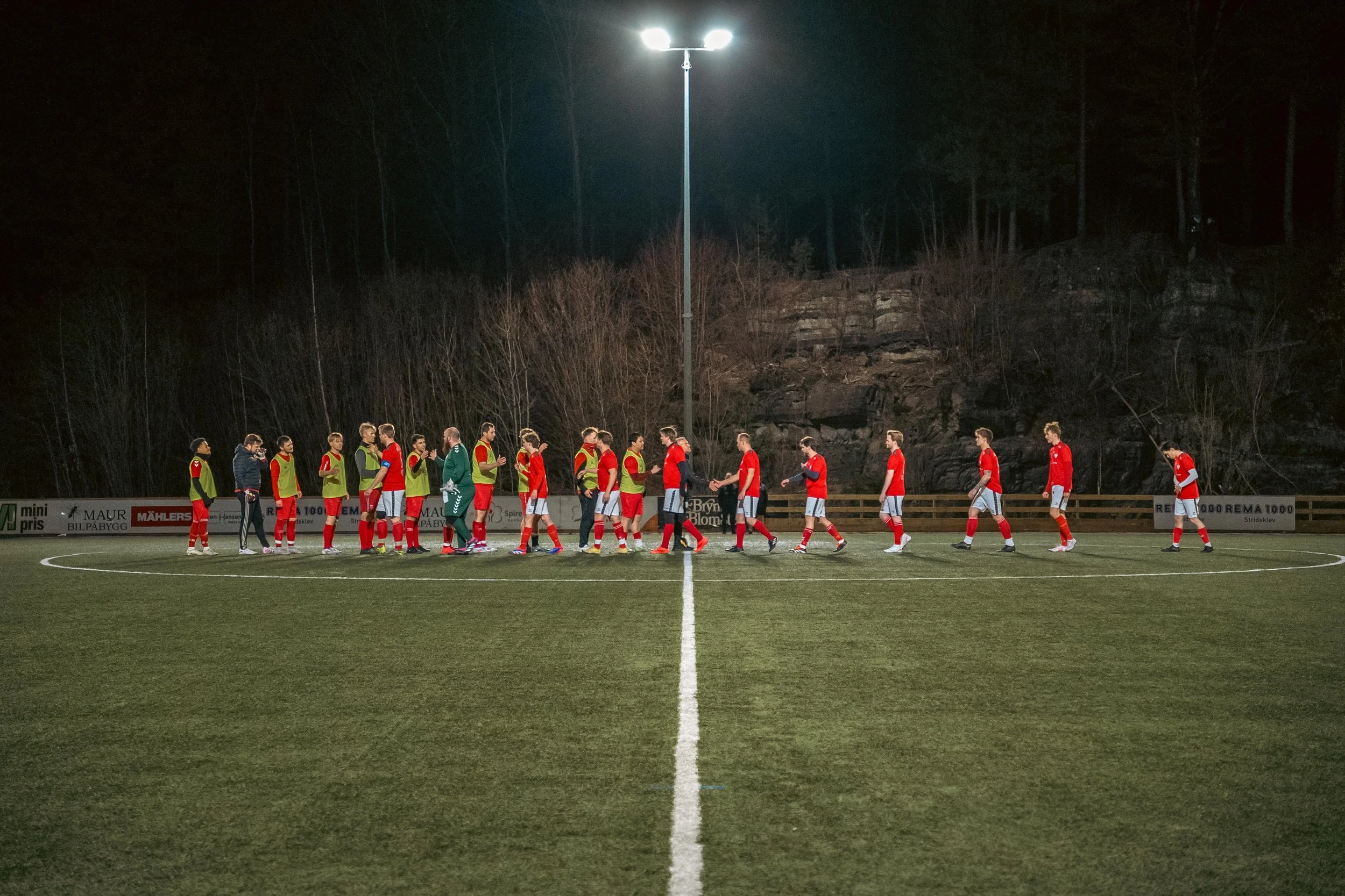 A soccer team in red jerseys and white shorts after a match, shaking hands with a team in green training bibs, under bright stadium lights at night.
