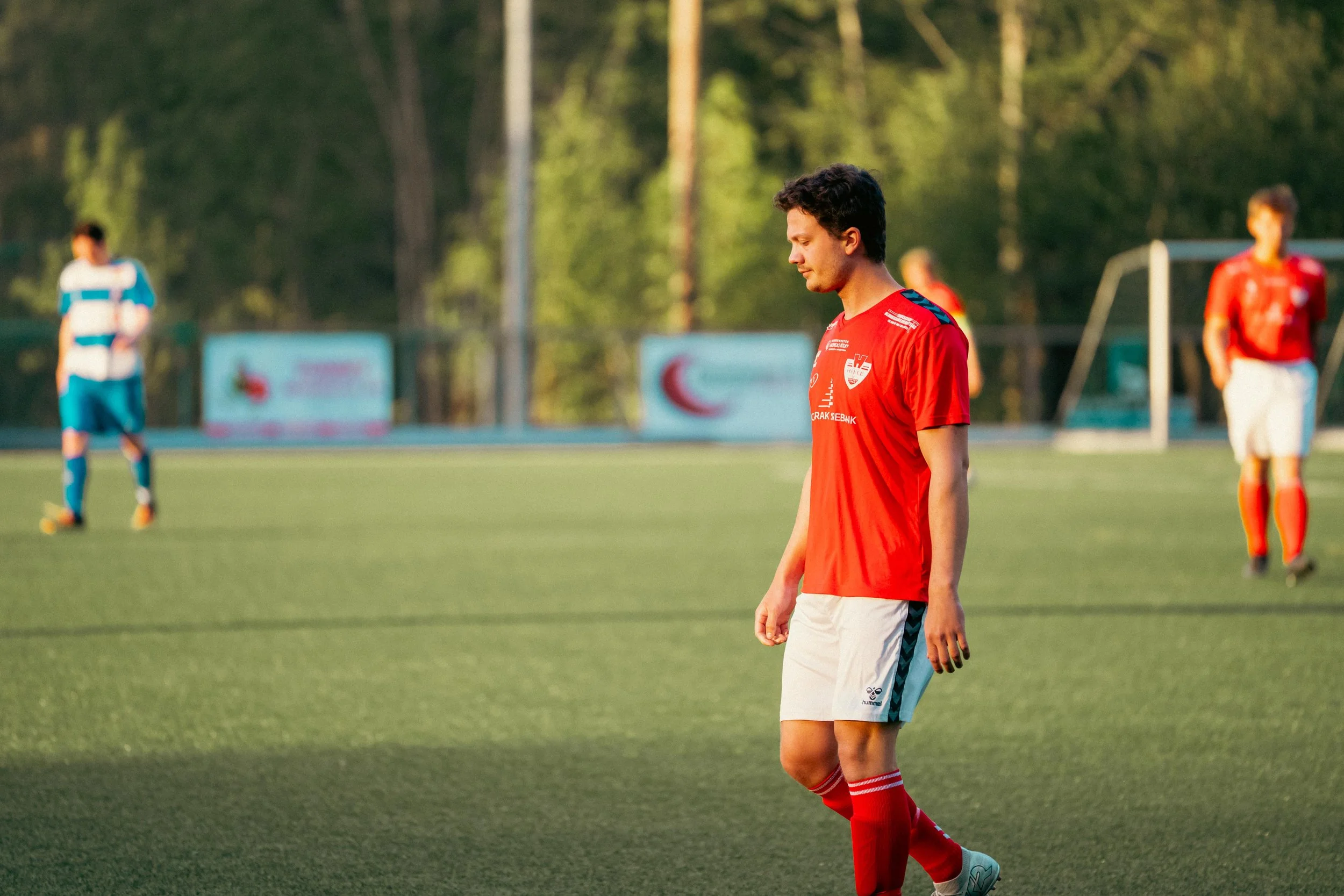 A soccer player in a red jersey, white shorts, and red socks standing on a soccer field with other players in the background.