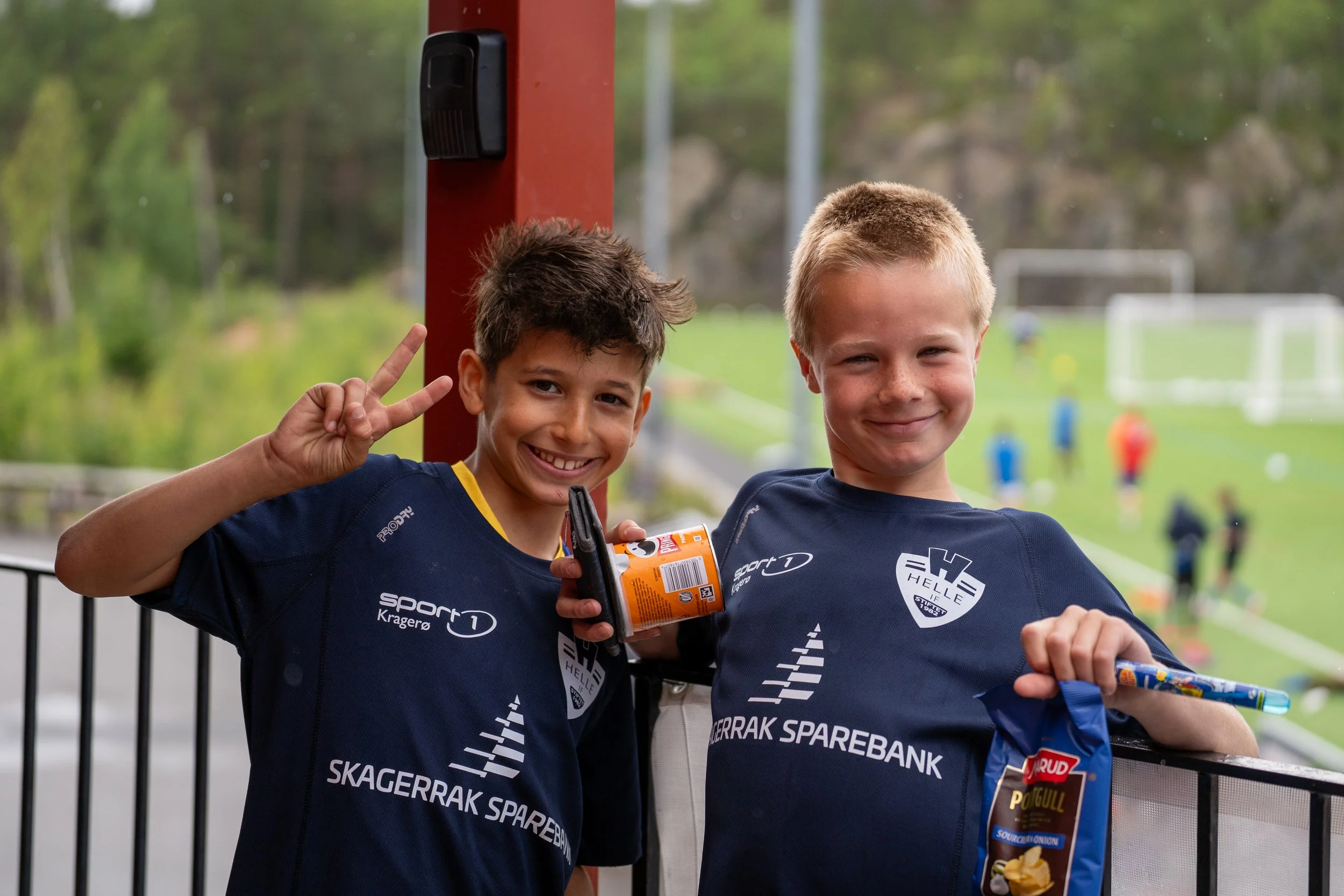 Two young boys wearing blue sports jerseys with sponsorship logos, smiling and posing for the camera at a sports field. One boy is holding a bag of potato chips and a snack, while the other is making a peace sign with his hand.